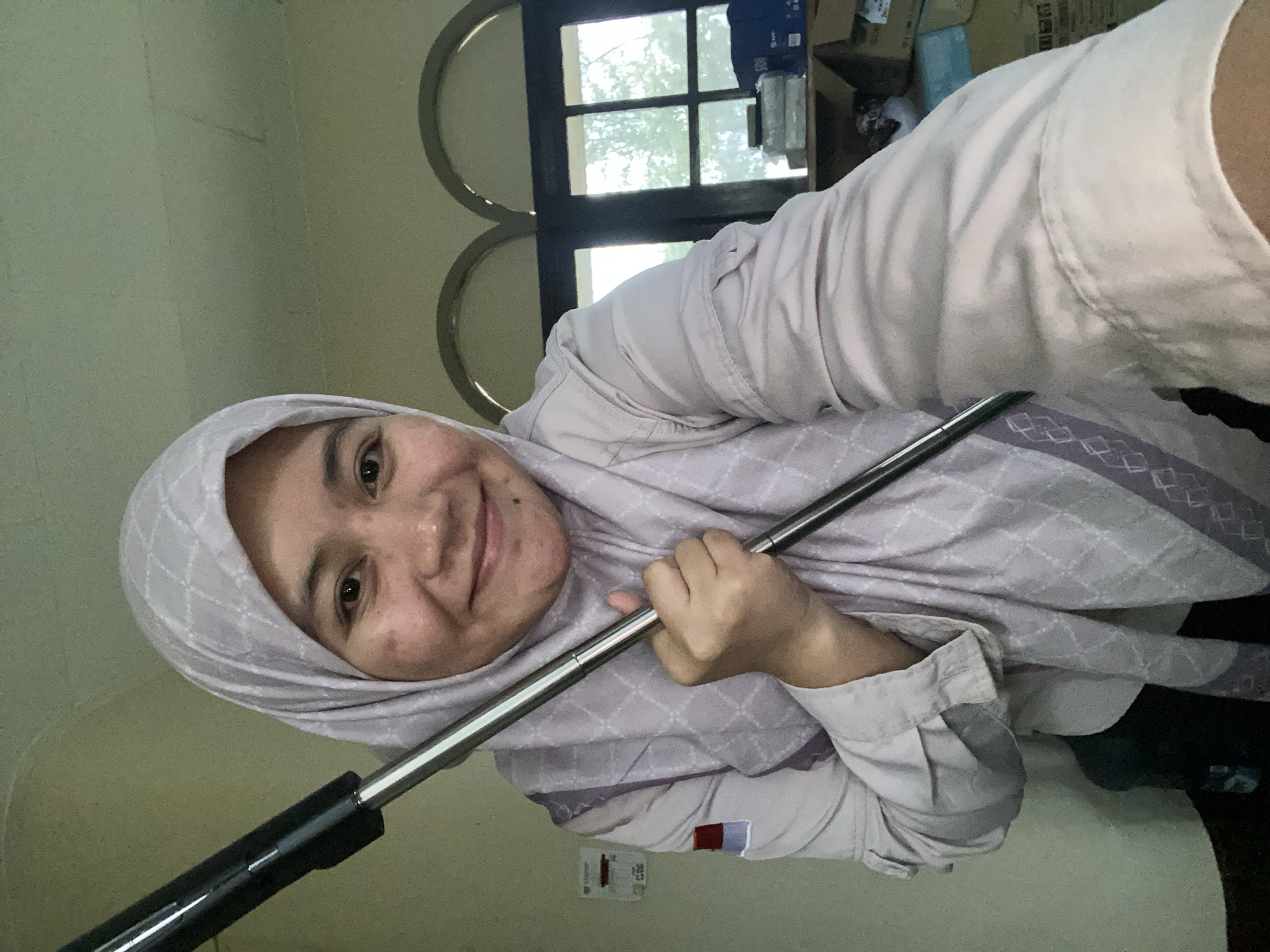 In a selfie taken indoors, a young woman wearing a light purple hijab and a light purple collared shirt is smiling at the camera. She is holding a mop handle with her right hand, the metallic handle extending upwards and to the left of the frame. The background appears to be a room with pale green walls and a window with arched panes. Behind her, to the right, shelves are visible with boxes and other items. The woman's skin has some blemishes. The image is captured at a close range, with the woman's face and upper body filling most of the frame. Based on the provided context, this scene is located in Banjarbaru, Indonesia.