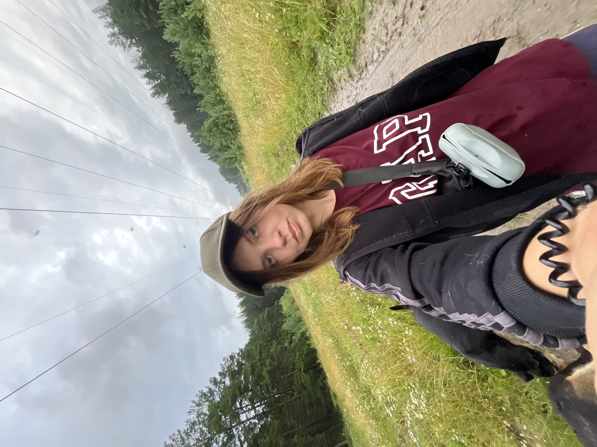 A young Caucasian female wearing a green baseball cap, a maroon t-shirt with white lettering, and a black jacket is captured in a selfie. She is walking on a muddy path through a grassy field dotted with small white flowers. A dark-colored dog is partially visible in the lower left quadrant of the image, walking alongside her. Power lines are visible overhead, stretching across a cloudy sky. The background features a dense forest. The scene appears to be outdoors, possibly in a rural or park-like setting, consistent with areas found in Nizhny Tagil, Russia.