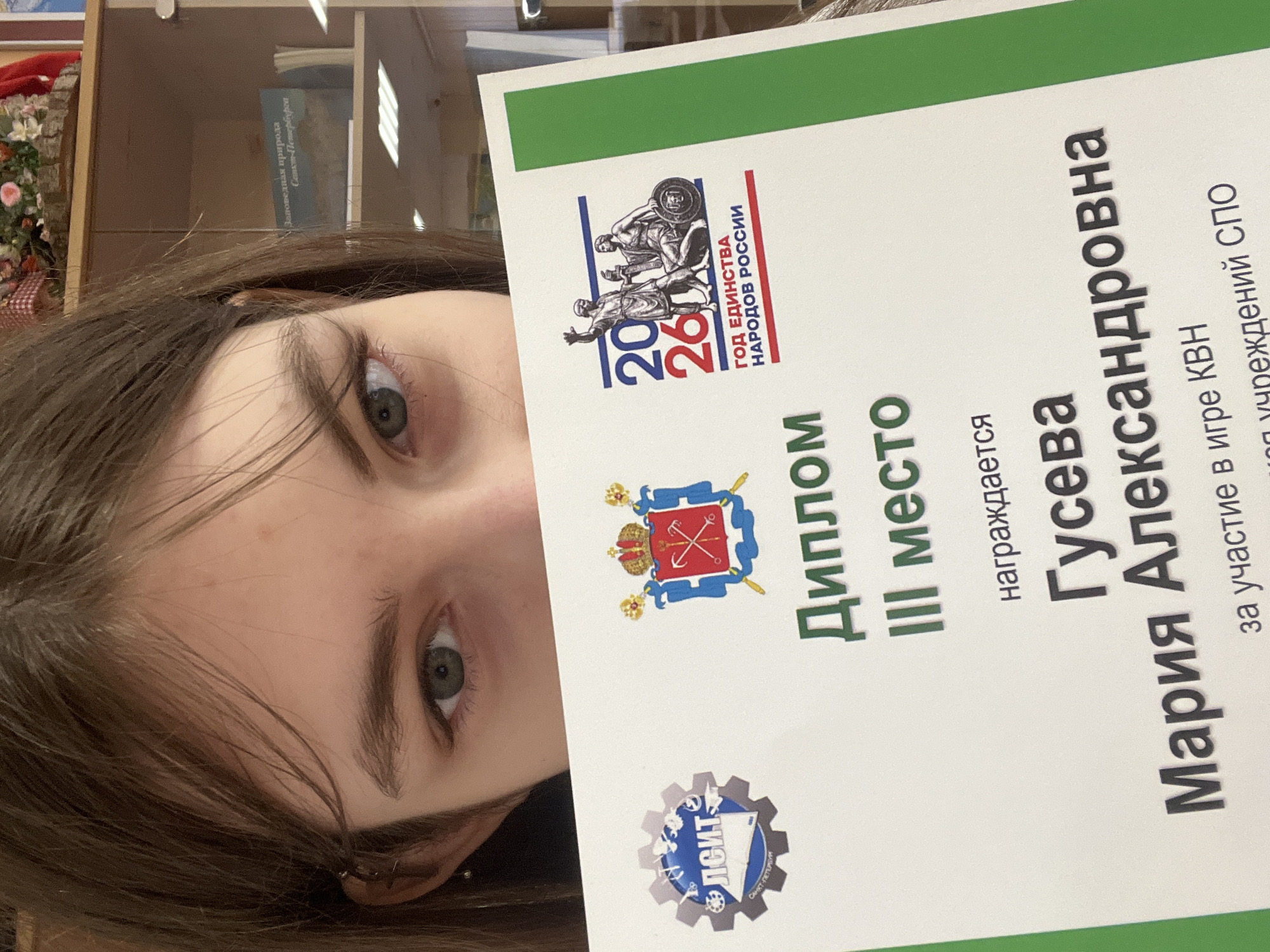 A young woman with dark hair and light eyes is holding up a certificate that reads, "Diploma, 3rd Place" and has the name "Maria Alexandrovna Guseva." The certificate also mentions "Participation in the KVN game" and the abbreviation "SPO" as well as the year "2026" and "the year of the Unity of the peoples of Russia." The coat of arms of the city of Saint-Petersburg can be seen on the certificate. The abbreviation ЛСИТ (LSIT) and "Saint-Petersburg" are printed at the bottom. A bookcase with books is visible in the background.