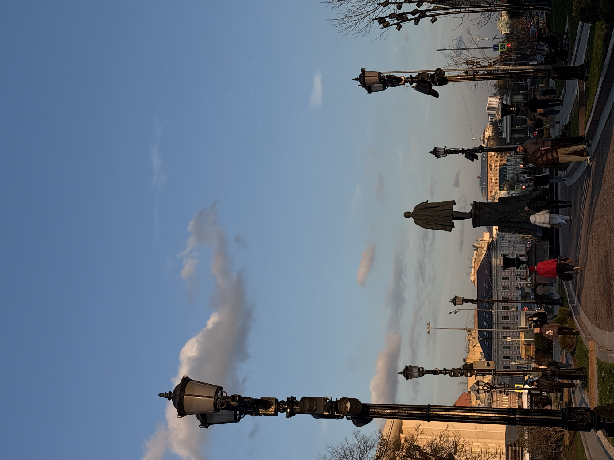 This image depicts a street scene in Moscow, Russia. The perspective is oriented lengthwise, with a statue of a person visible in the middle ground, standing on a pedestal. Pedestrians are walking along the sidewalk next to the statue. Street lamps, with a distinctive design, are spaced evenly along the pavement. The background features buildings and trees. The sky is blue and contains some clouds.