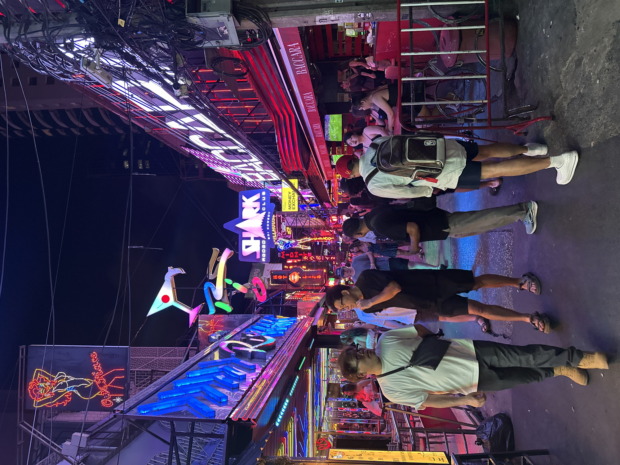 The image shows a street scene at night in Bangkok, Thailand. The street is brightly illuminated with numerous neon signs for bars and clubs, including "SHARK CLUB AGO GO", "BACCARA", and "SOI COWBOY". A crowd of people is walking along the street. The street is crowded with power lines and cables overhead. The ground is paved and has some trash.