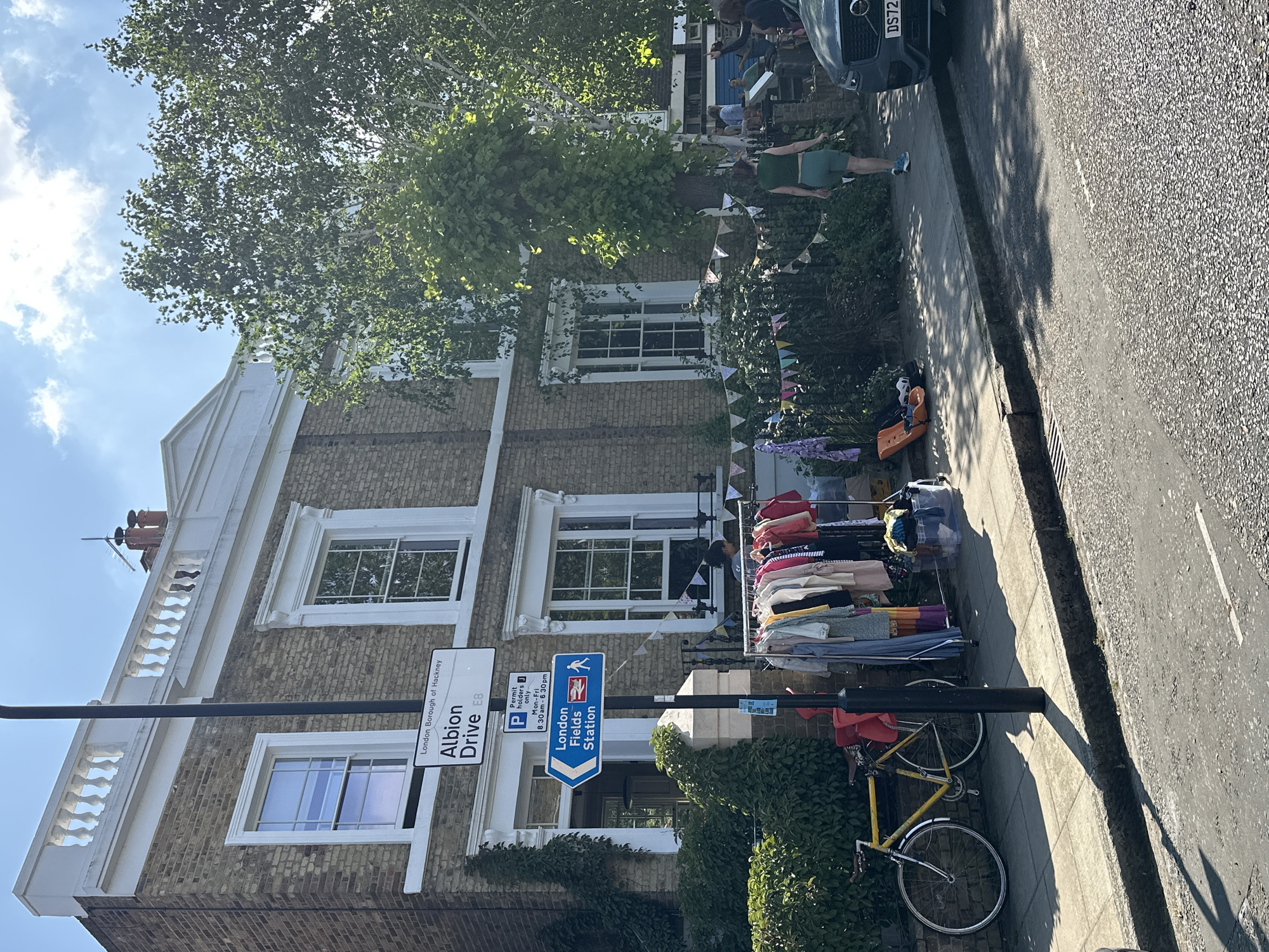 The image shows a residential street in the London Borough of Hackney, specifically Albion Drive, E8. The building on the left is a three-story brick house with white window frames and a white railing on the roof. There is a street sign indicating "Albion Drive E8" and a parking permit sign. Adjacent is a sign pointing towards "London Fields Station" and indicating that the station is a short walk away. A clothes rack full of clothes is placed on the sidewalk in front of the house. A yellow bicycle leans against the pole of the street sign. A person is walking on the sidewalk further down the street, and part of a car is visible on the right side of the image. Bunting hangs from the trees and building behind the rack of clothes.