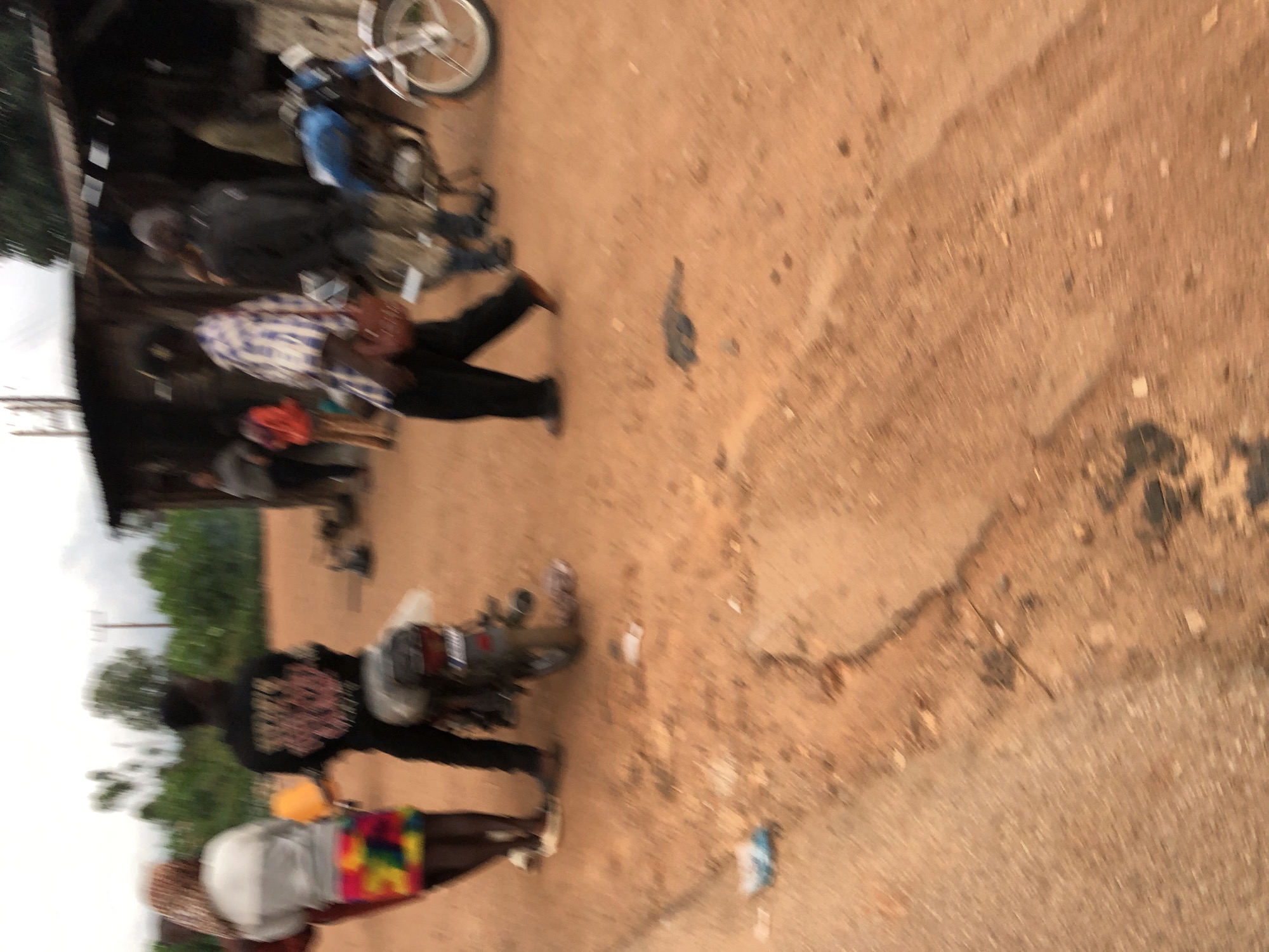In Vom, Nigeria, a dirt road scene unfolds with several individuals and a motorcycle. On the left, a person wearing a headscarf and a white shirt stands beside a colorful patterned skirt and dark leggings. Next to them, another person in a black shirt with pink lettering is visible. A motorcycle is parked between them. Further into the scene, a person in a plaid shirt and dark pants walks away from the camera. Behind them, two individuals are partially obscured, one in a black jacket and the other in a patterned shirt. To the right, a blue motorcycle is parked. In the background, a rustic structure with a corrugated metal roof stands. The ground is a mixture of reddish-brown dirt with scattered debris. The overall atmosphere is that of a rural or semi-urban street.