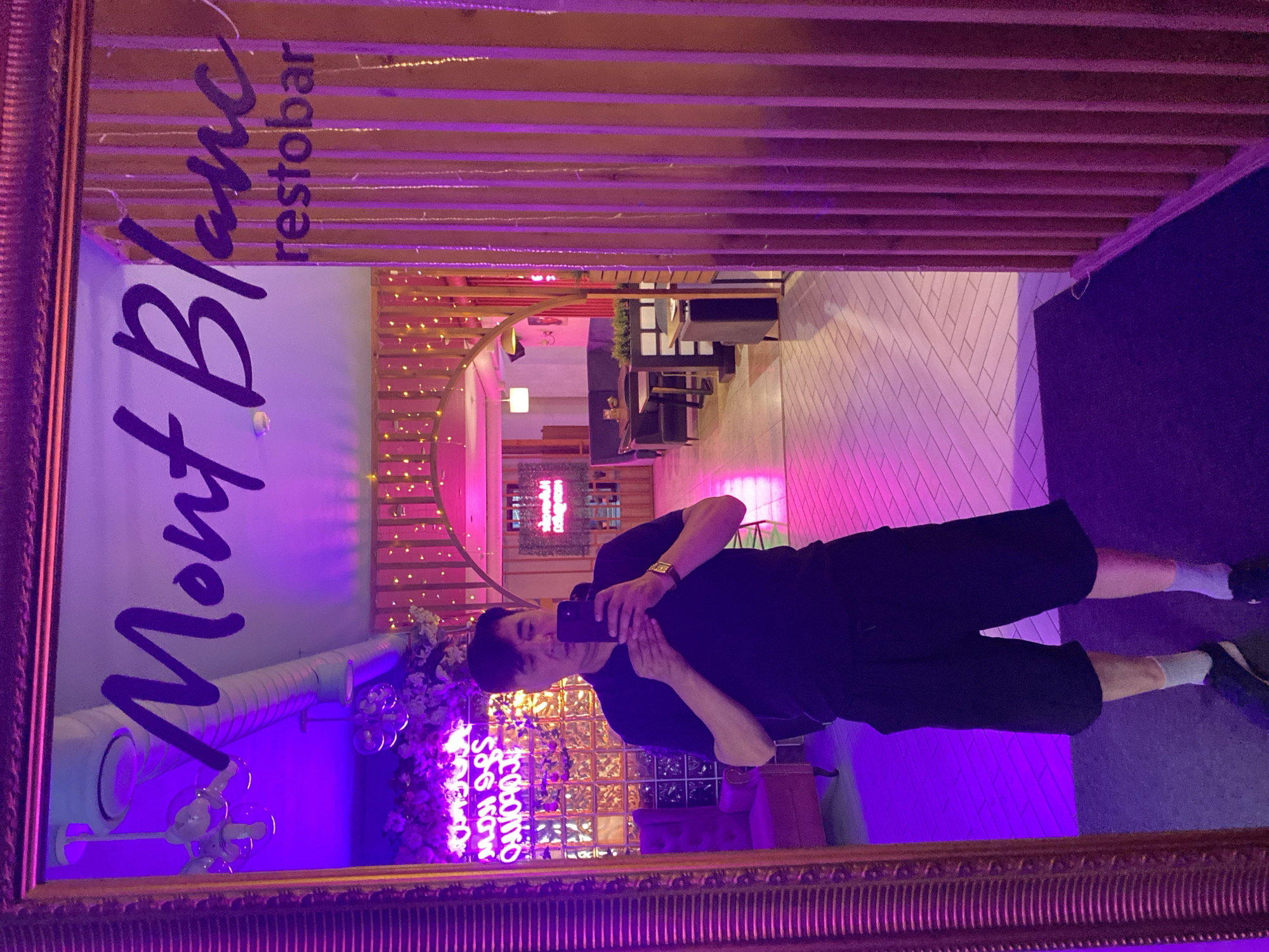 A man is taking a selfie in a mirror at Mont Blanc restobar in Petropavl, Kazakhstan. The man is wearing a black t-shirt, black shorts, grey socks, and black sneakers. He is holding a black smartphone in his right hand. The restobar is decorated with neon lights and string lights. The lighting is predominantly purple and pink, creating a vibrant and modern atmosphere. In the background, tables and seating areas are visible, suggesting a dining or bar setting. The mirror is large and ornate, with a gilded frame.