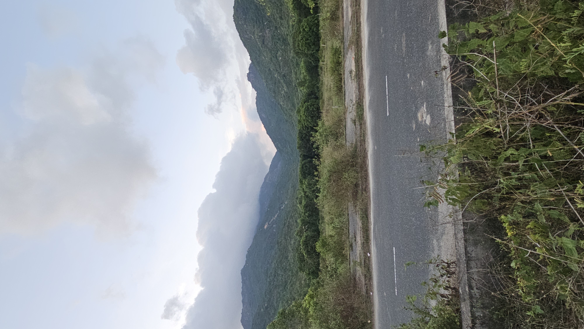 The image depicts a road in Cam Lam, Vietnam. The road runs along the edge of a mountain range covered in green vegetation. Clouds partially obscure the mountain peaks. The road is gray, with white lane markings. A variety of weeds and shrubs grow along the road's edge. The sky is overcast.