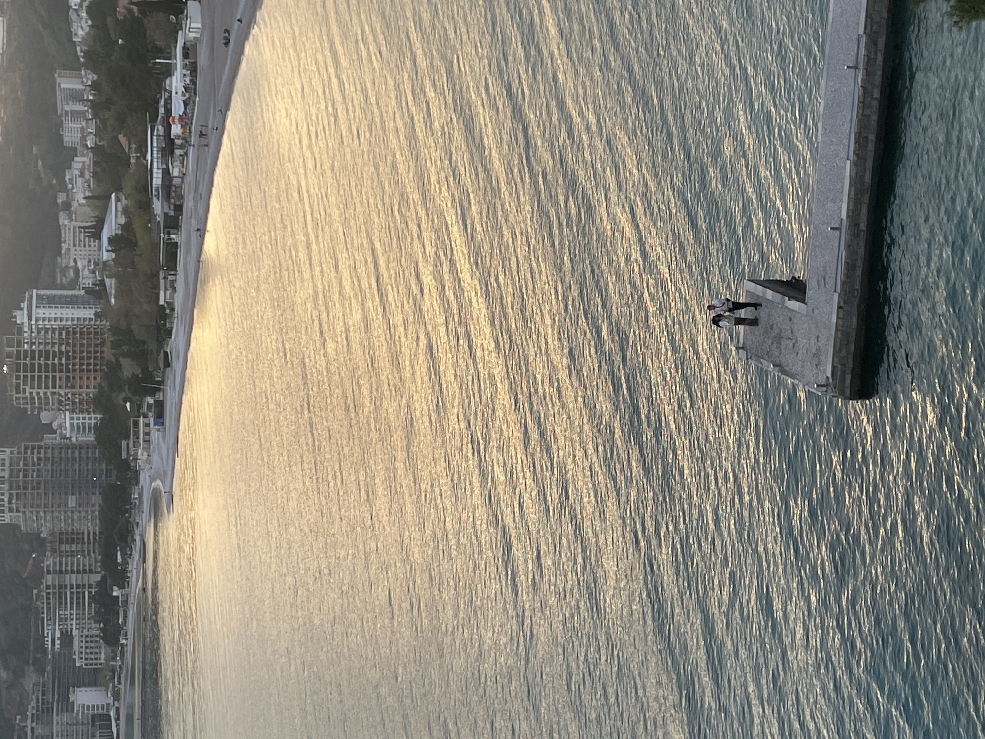 The image shows a high-angle view of a pier extending into the sea, with the city of Budva, Montenegro, visible along the coast. Two people stand at the end of the pier. The sea reflects the sunlight, creating a shimmering effect. The city is characterized by buildings of varying heights and architectural styles.