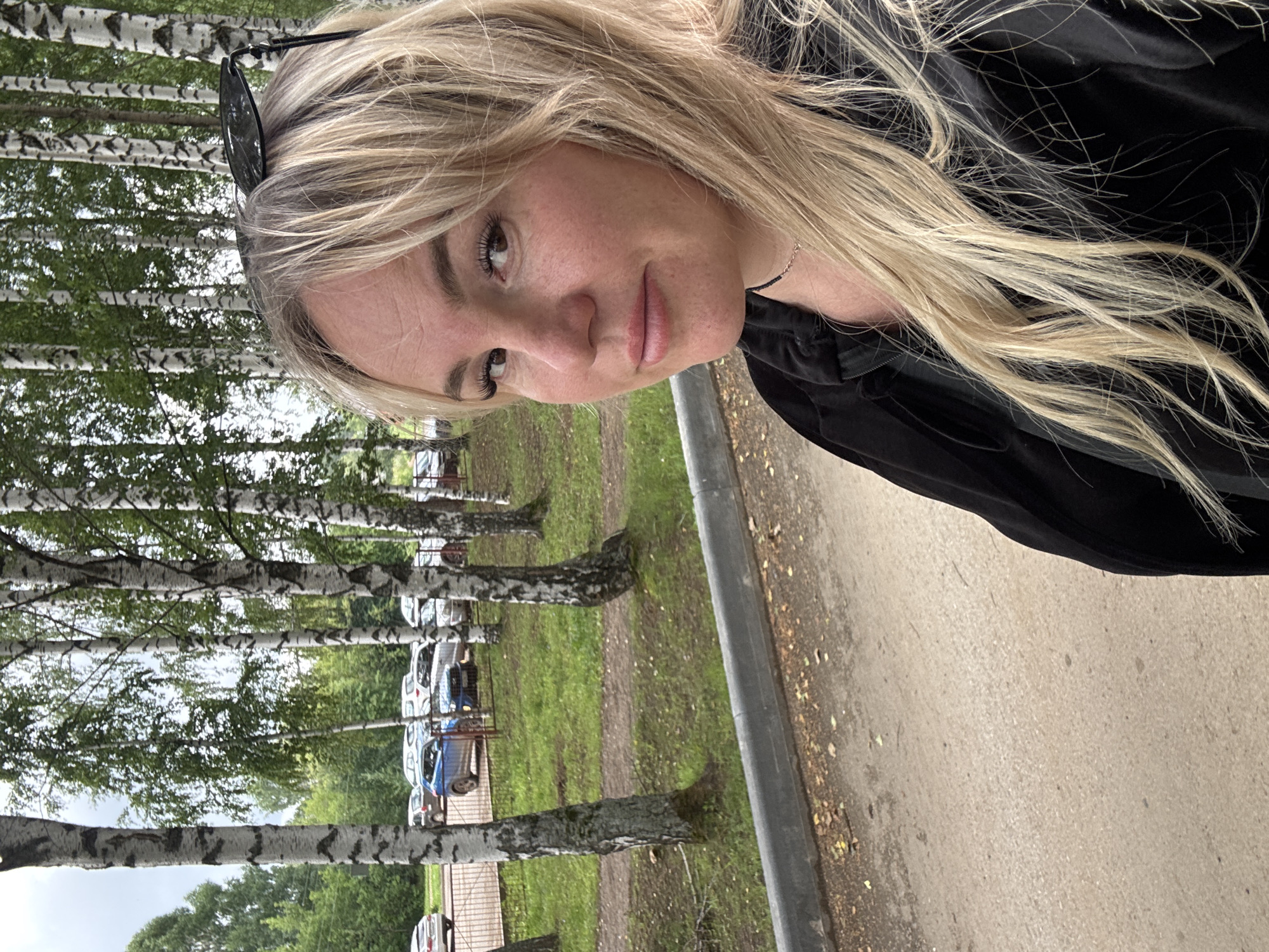 A medium shot captures a blonde woman with fair skin, wearing a black jacket and a thin silver necklace. She has long, wavy blonde hair and is looking towards the camera with a slight smile. Sunglasses are perched on top of her head. She is outdoors, with a row of birch trees behind her. In the background, several parked cars are visible in a lot adjacent to a grassy area. The ground in the foreground is a dirt path, bordered by a grey concrete curb. The scene appears to be in a park or residential area in the city of Chastyye, Russia.