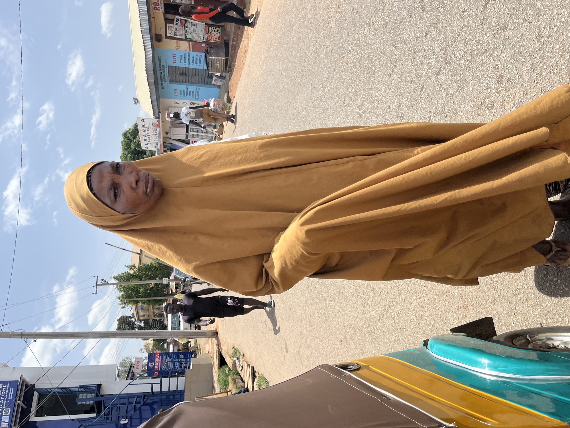 The image shows a woman in a golden-brown hijab walking along a street in Jos, Nigeria. The street scene includes several businesses with signs, including a medical diagnostic lab ("ELYON MEDICAL DIAGNOSTIC LABORATORIES") that lists tests like typhoid, genotype, blood group, malaria, hepatitis, and diabetes, suggesting a healthcare-related context. Another business advertises printing and photocopy services ("PRINTING PHOTOCOPY POS"). In the background, there are people walking and signs for other businesses like "BARKA COSMETICS" and "COMMUNICATIONS PHONES & ACCESSORIES". There is also a vehicle with a yellow and teal color scheme. The sky is partly cloudy.