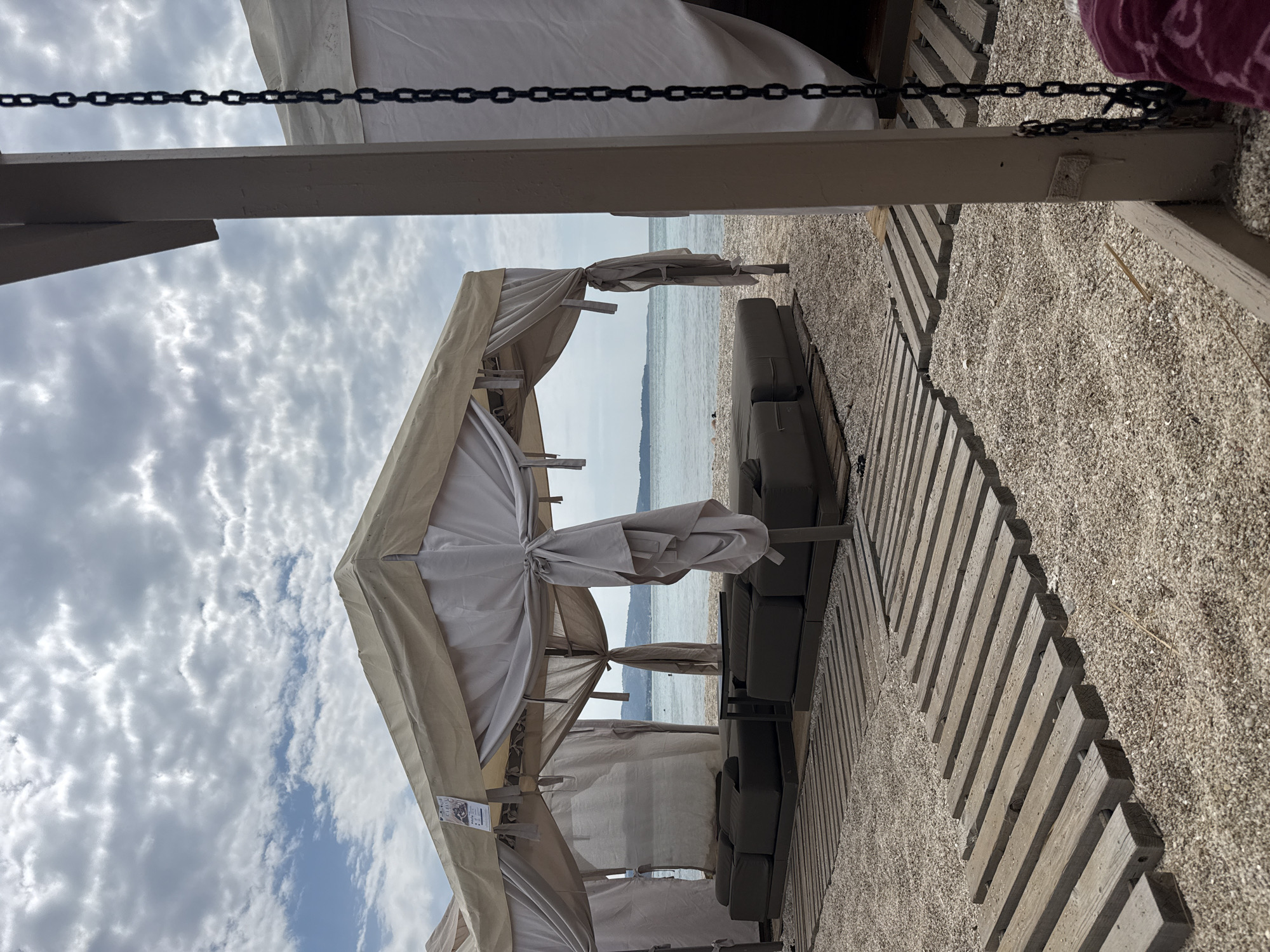 A beach scene in Limenaria, Greece, features a covered seating area on a pebble beach. The structure consists of a tan canopy supported by poles, with drapes of light fabric. Beneath the canopy are several dark-colored modular lounge seats. Wooden plank pathways are laid on the pebble surface, leading towards the seating area and extending into the background. Beyond the beach, a body of water is visible, with a distant coastline featuring hills. The sky is overcast with scattered clouds. A metal chain hangs from a wooden beam on the right side of the frame, partially obscuring a white fabric structure. The scene appears to be set up for relaxation or a beachside lounge.