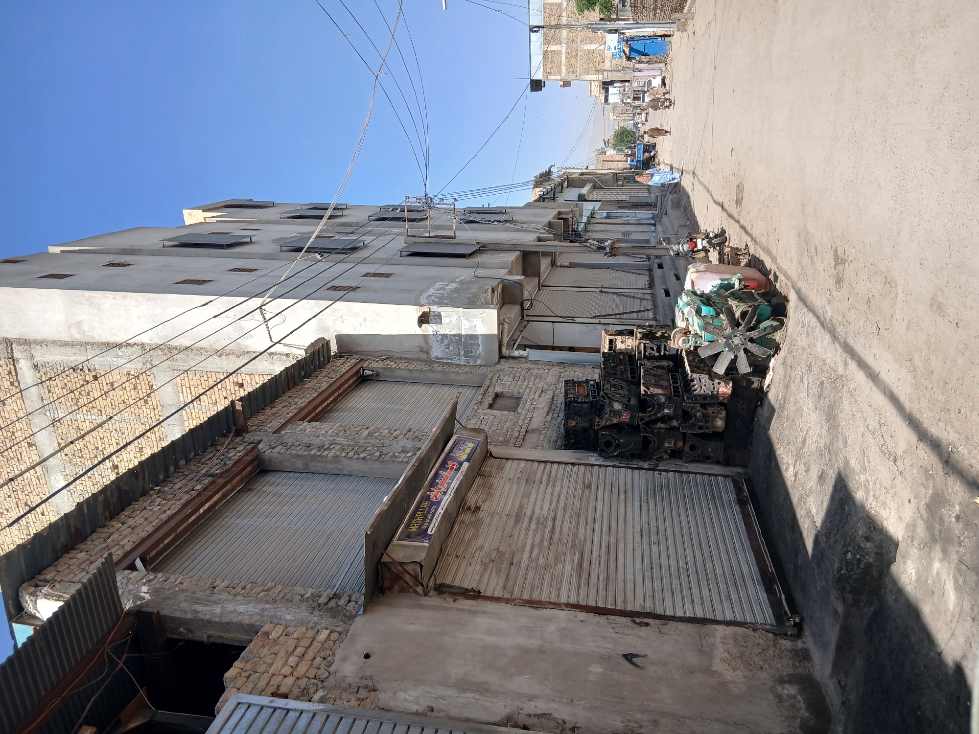 The image depicts a street scene in Quetta City Tehsil, Pakistan. The view is angled upwards, showing a row of aging buildings with closed metal shutters on the ground level storefronts. The buildings appear to be constructed from a combination of brick and concrete, showing signs of wear and disrepair. Power lines stretch across the sky above the buildings. Adjacent to the buildings on the right side of the frame, there is a collection of discarded engine blocks and a teal-colored mechanical fan. The street extends into the distance, revealing other buildings and a few pedestrians or animals farther down the road. The sky is clear and blue.