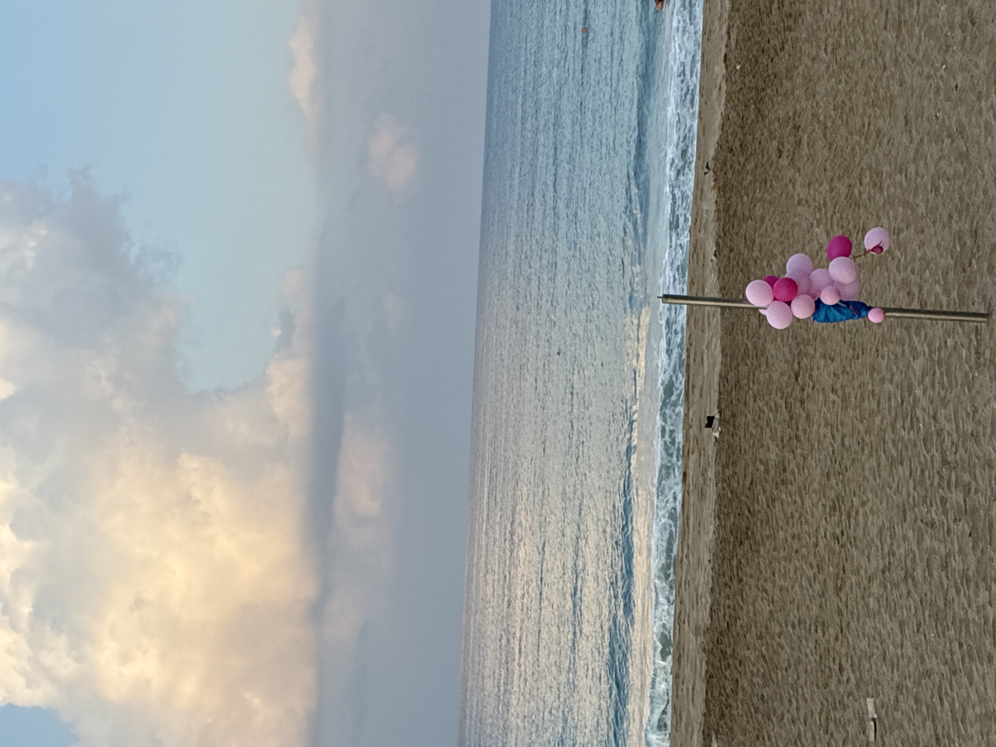 This image depicts a beach scene in Alanya, Turkey. The foreground shows sandy beach with a metal pole to which several pink balloons are attached. A blue piece of fabric is also tied to the pole. In the midground, gentle waves lap onto the shore. The background is dominated by the sea, with a clear horizon line separating it from a sky filled with scattered clouds, some tinged with the warm hues of sunrise or sunset. The overall impression is one of a calm, possibly early morning or late evening, beach setting.