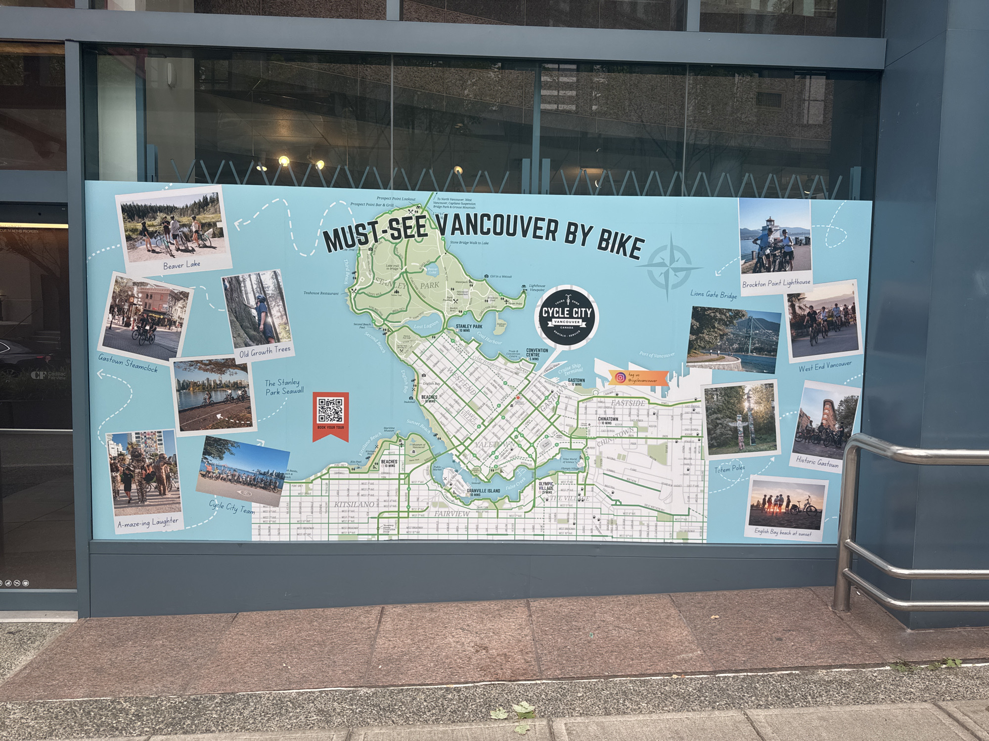 A large, light blue map titled "MUST-SEE VANCOUVER BY BIKE" is displayed on an exterior wall, likely outside a building in Vancouver, Canada. The map features a detailed illustration of Vancouver's downtown peninsula and Stanley Park, with marked bicycle routes indicated by green lines. Numerous photographs are placed around the map, each accompanied by a caption and depicting various locations and activities in Vancouver.
Specific locations highlighted in the photographs include:
- Beaver Lake
- Gas town Steamclock
- Old Growth Trees
- The Stanley Park Seawall
- Brockton Point Lighthouse
- Lions Gate Bridge
- West End Vancouver
- A-maze-ing Laughter
- Cycle City Team
- Totem Poles
- Historic Gastown
- English Bay beach at sunset
The map also includes a QR code for booking a tour and social media handles. The overall display appears to be an advertisement or informational piece for cycling tourism in Vancouver. The environment is urban, with the map mounted on a building with a reflective glass facade.
