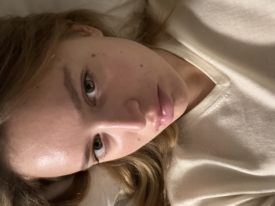 A fair-skinned young woman with light brown hair reclines, gazing upward. Her eyes are hazel, and she has full lips with a glossy sheen. Freckles dot her cheek and forehead. She wears a creamy, light-colored shirt with a visible neckline. The image is softly lit, accentuating the contours of her face.