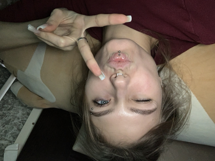 The image displays a close-up, chest-up self-portrait of a female subject, observed indoors in the city of Фоки, Russia. The subject has light brown hair, fair complexion, and blue irises. Her left eye is closed in a wink, while her right eye remains open. Facial piercings include a silver horseshoe-style septum ring, two silver vertical labret studs on the lower lip, and two silver studs piercing the right eyebrow. The subject's lips are pursed. Her left hand is raised, with the thumb and index finger extended to form an 'L' shape, and the remaining fingers curled; a silver ring is present on the ring finger. Her fingernails are long and feature white tips. She wears a dark red hooded garment. The background is dimly lit. To the subject's left, a light-colored pillow and an orange-brown object are partially visible. To the right, behind the subject's upper right arm, an irregularly shaped, light-tan object is present, partially obscured by two parallel white adhesive strips.