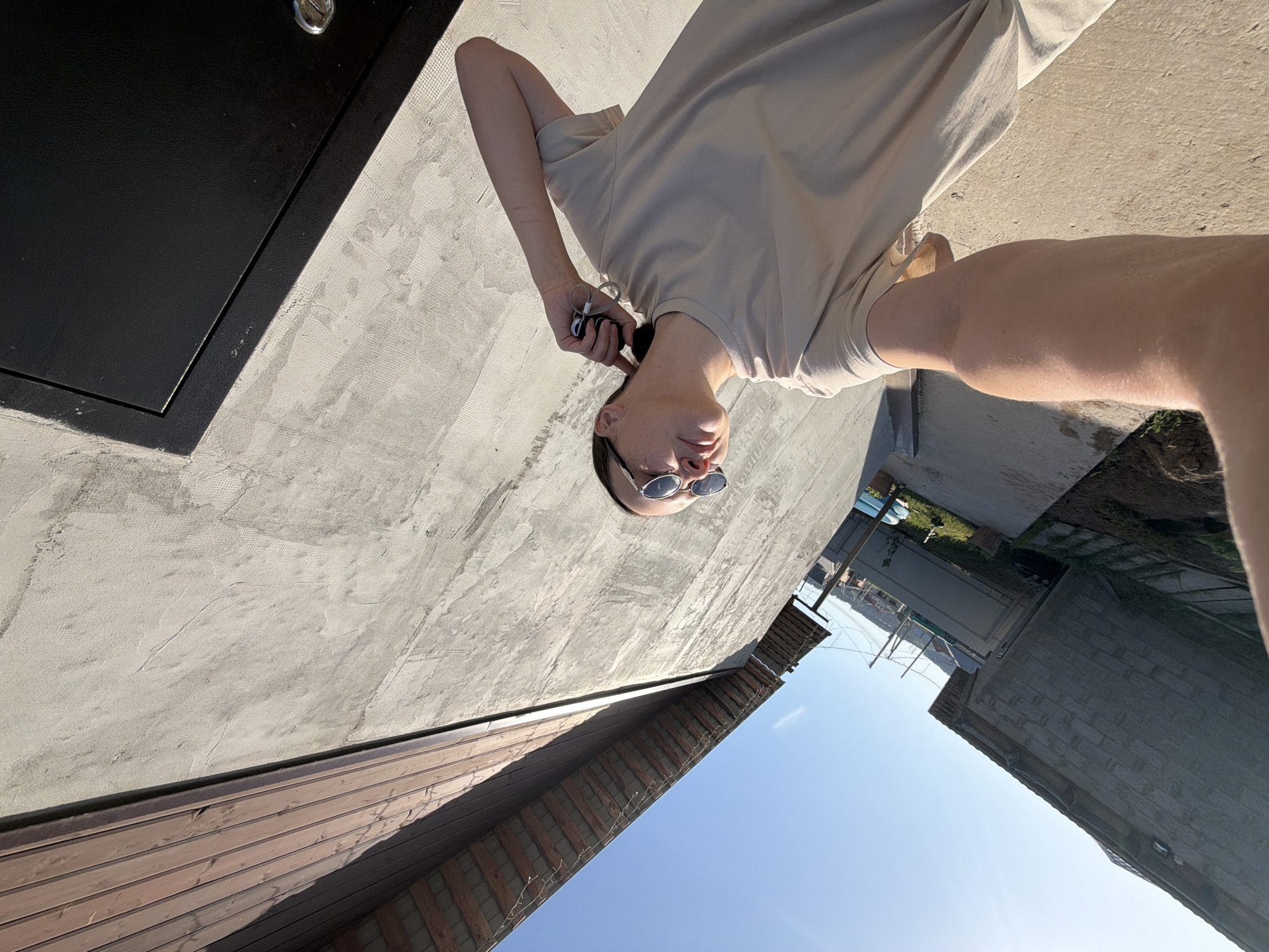A medium-angle outdoor shot captures a person from the mid-thigh up, clad in a beige t-shirt and sunglasses, with keys dangling from their left hand. The person is situated on a concrete surface. To the left, there's a dark, flush-mounted box set into a textured concrete wall. A wooden facade runs along the base of the wall. In the background, a residential area is visible under a clear blue sky, suggesting the location is in Chaikovsky, Russia.