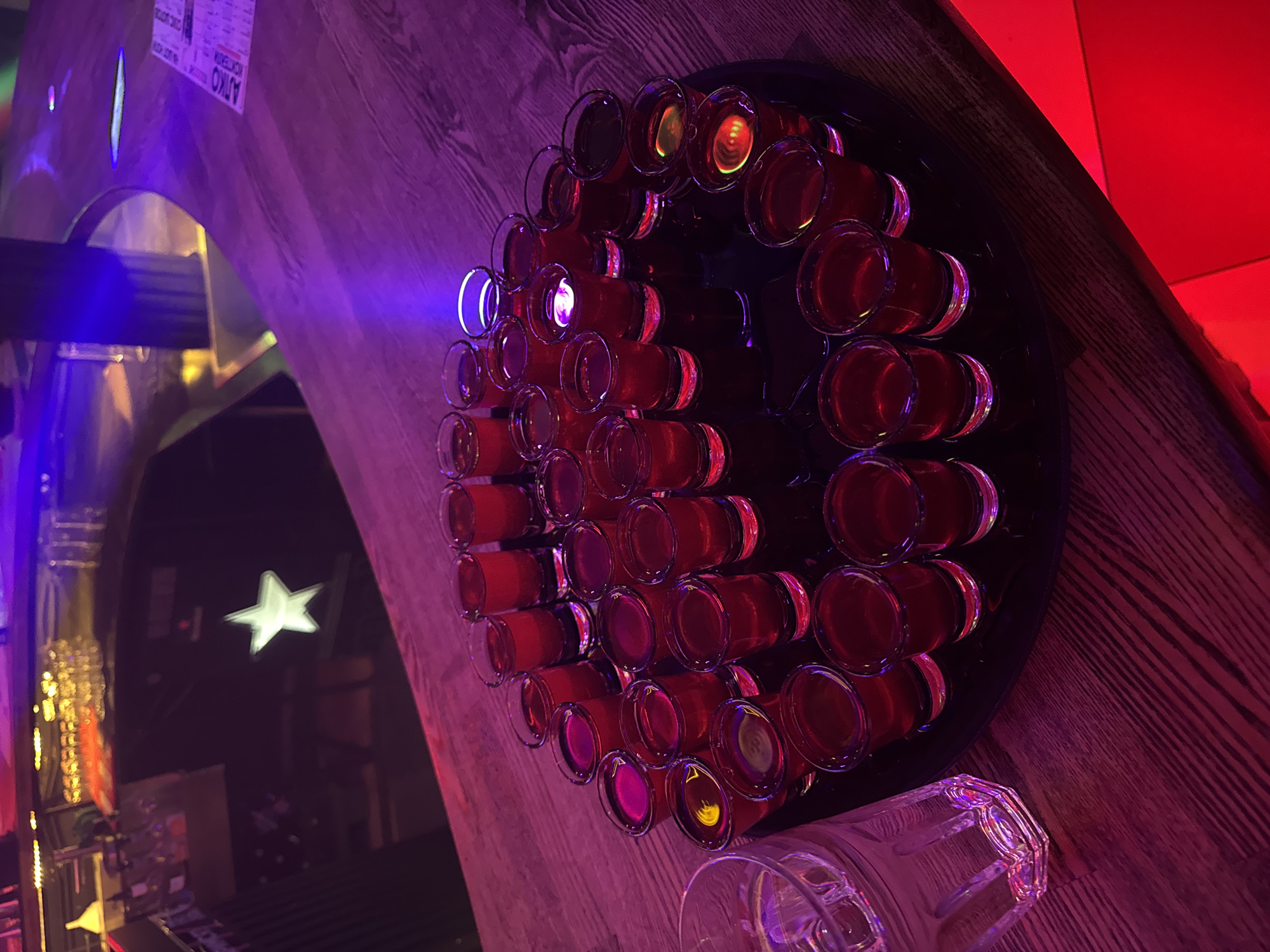 The image shows a collection of red shots arranged on a dark tray. The tray itself is placed on a wooden surface. The lighting in the scene is dominated by purple and red hues, creating a dim and atmospheric setting, likely a bar or nightclub. There are some blurry elements in the background, including what appears to be a white star-shaped light. There are also some labels visible on a wooden surface in the upper left corner of the image, but the text is illegible. The overall impression is of a lively evening or night out.