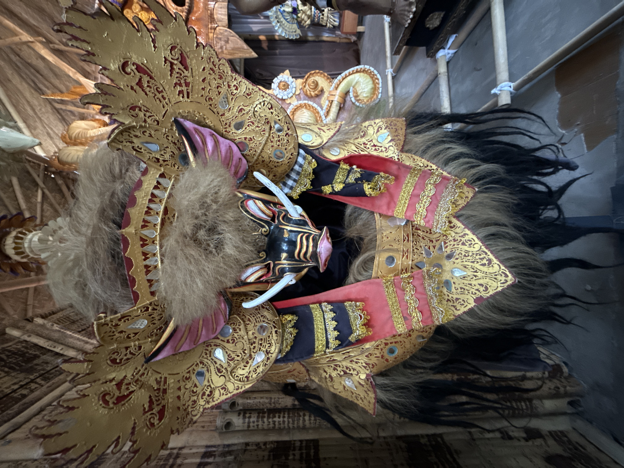 A close-up, head-on shot depicts a large, elaborate mask on display, likely part of a traditional Balinese performance or art installation. The mask, characterized by a stylized boar's face with prominent tusks, is adorned with intricate gold leaf detailing, vibrant red and black fabric accents, and a voluminous mane of brown and black hair. Large, golden, wing-like structures, also embellished with gold patterns, extend from the sides of the mask. The mask is set against a backdrop of bamboo-like material, suggesting an outdoor or rustic setting. In the background, other decorative elements and structures, including what appear to be other masks or figurines, are visible. The scene is lit with ambient light, creating some shadows and highlighting the textures and metallic sheen of the mask. The overall impression is one of cultural artistry and dramatic presentation. The location is Kuta Selatan, Indonesia.