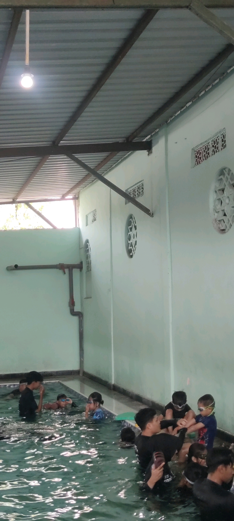 In Gilang, Indonesia, a group of children and an adult are present in an indoor swimming pool. The pool has a shallow area and is surrounded by a tiled edge. The walls of the building are painted a light green color, and there are several decorative circular and rectangular vents on the wall. The ceiling is made of corrugated metal panels supported by metal beams, and a bare light bulb hangs from the ceiling. In the water, the children are wearing swimsuits and goggles. Some are actively swimming, while others are interacting with each other or the adult. The adult, a male, appears to be a swim instructor, assisting the children. The water is clear and reflects the light. There are pipes and fixtures visible on the wall near the edge of the pool, suggesting a water circulation system. The overall impression is a swimming lesson taking place in a covered outdoor or semi-indoor facility.