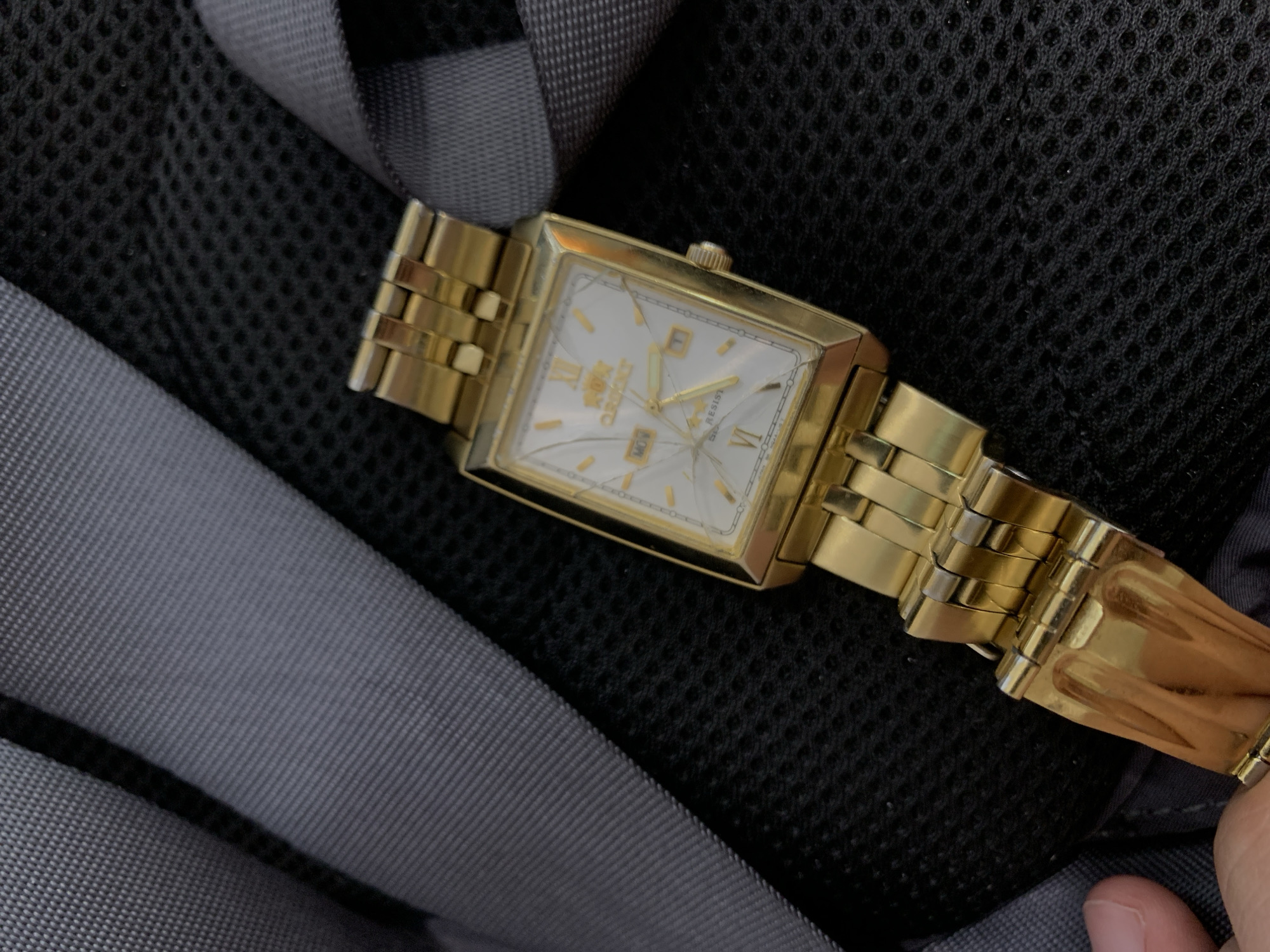 A gold-colored Orient wristwatch with a white face and Roman numerals is displayed on a black mesh fabric background. The watch has a day and date complication, with "MON" visible for the day and "11" visible for the date. The crystal face of the watch has several visible cracks. The watch band is also gold-colored and appears to be made of linked metal segments. The background is textured with a repeating hexagonal pattern, suggesting it could be part of a bag or other fabric item. A hand, partially visible on the left side of the image, is touching the watch band. The image is taken from a top-down perspective.