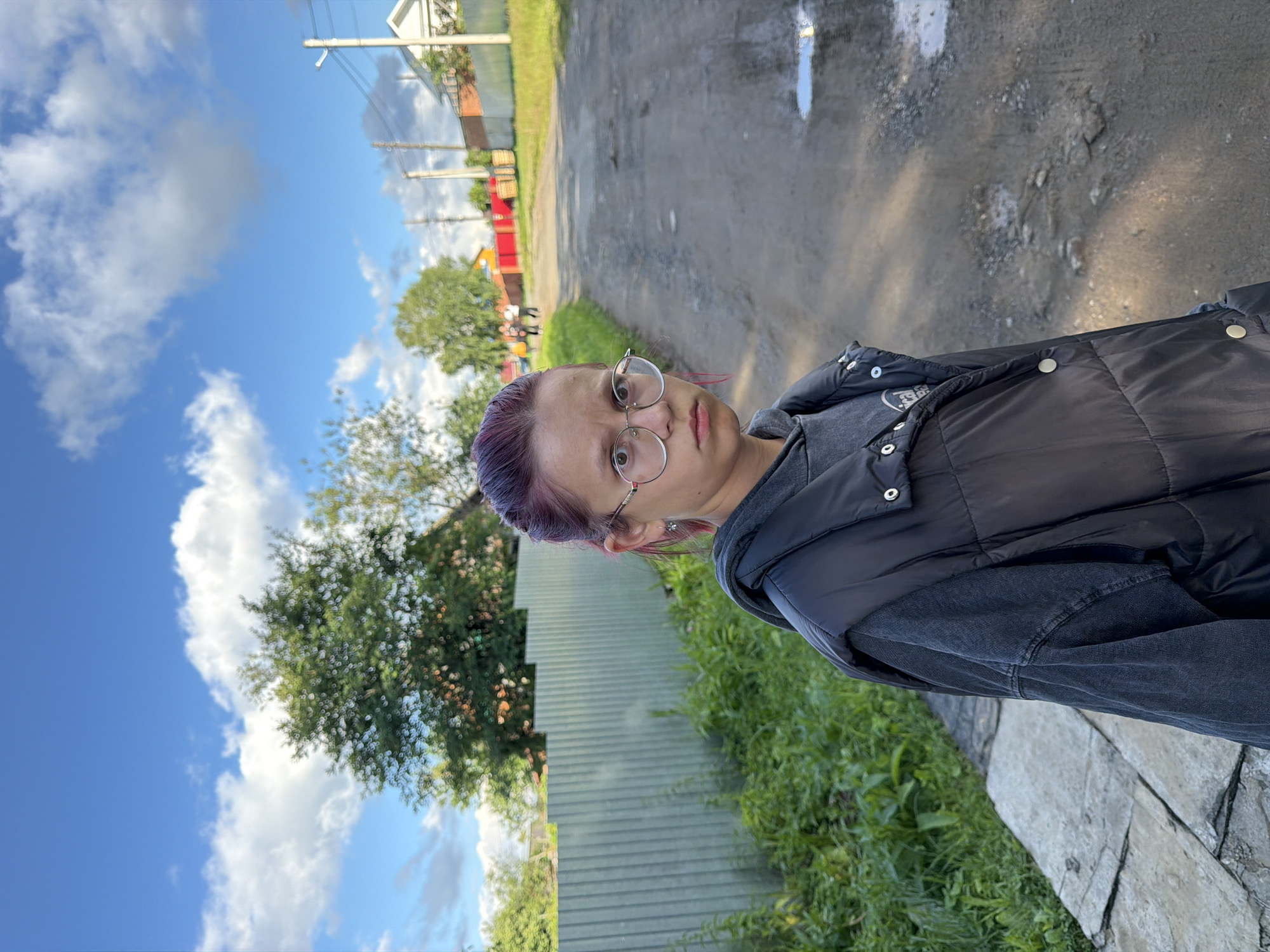 A medium shot captures a young person with dark hair, tinted pink at the ends, wearing a grey hoodie and a black puffer vest, standing outdoors in what appears to be a rural or suburban area. The person is looking directly at the camera with a neutral expression and wearing round, clear-rimmed glasses. To the left of the person is a tall, grey corrugated metal fence, bordered by lush green grass and foliage. Behind the fence, trees are visible. The ground in front of the person is a dirt path, with puddles of water indicating recent rain. In the background, a dirt road stretches into the distance, with houses and fences on either side. A few people can be seen walking in the background on the road. The sky is a vibrant blue with scattered white clouds. The scene is likely located in Chaikovsky, Russia, given the architecture and general environment.