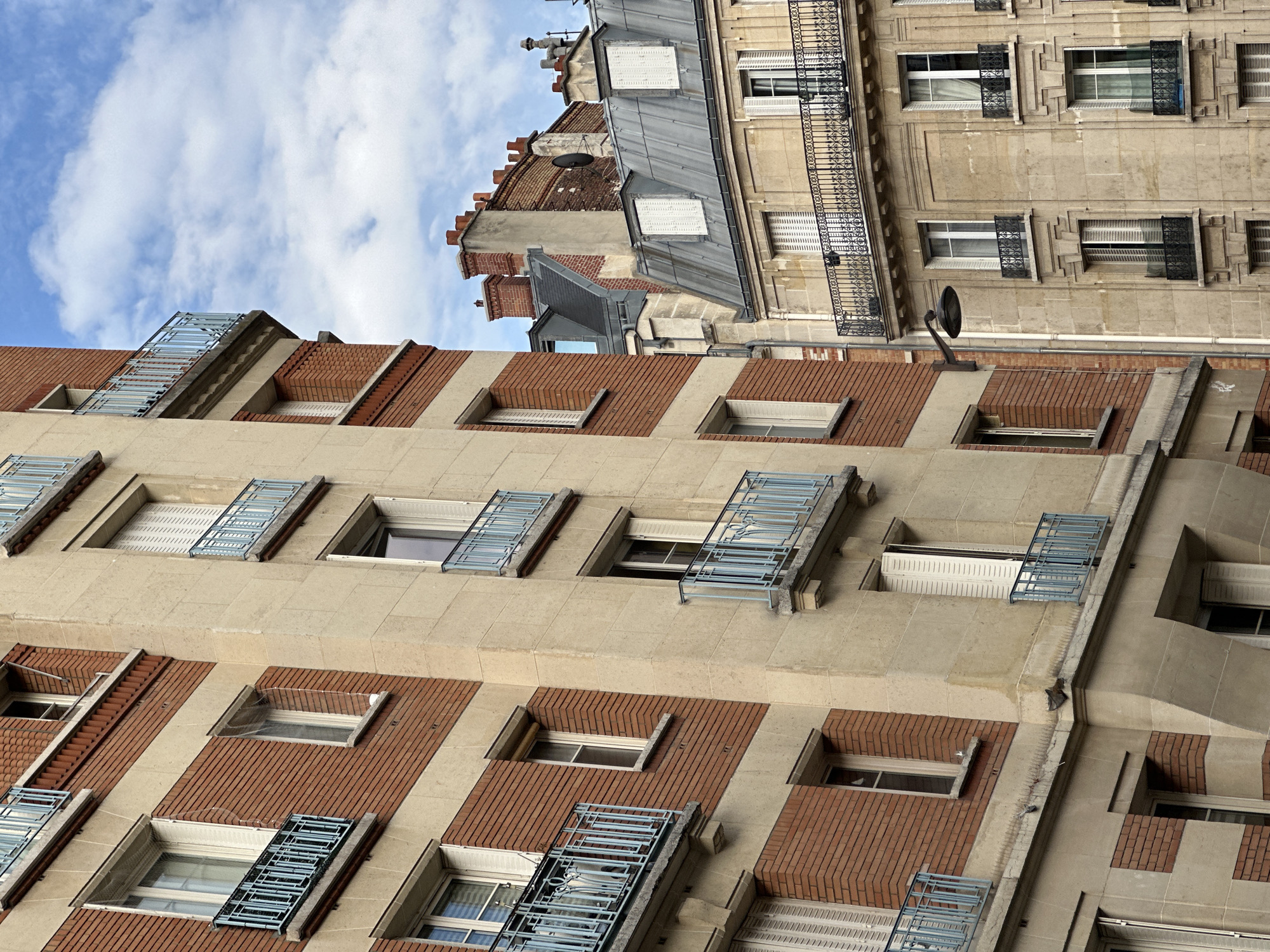 This is a medium-angle shot of Parisian buildings against a cloudy blue sky. The foreground features the facades of two buildings. The left building is characterized by alternating bands of red brick and beige stone. It has numerous windows, most with white shutters and some with light blue metal balconies. The right building, slightly further back and to the right, is constructed of beige stone blocks. It also features windows with white shutters and decorative wrought-iron balconies. Visible architectural elements include chimneys, gabled roofs, and drainpipes. A street lamp extends from the left building towards the right. The overall scene conveys an urban environment in Paris, France. No people are visible in the image.