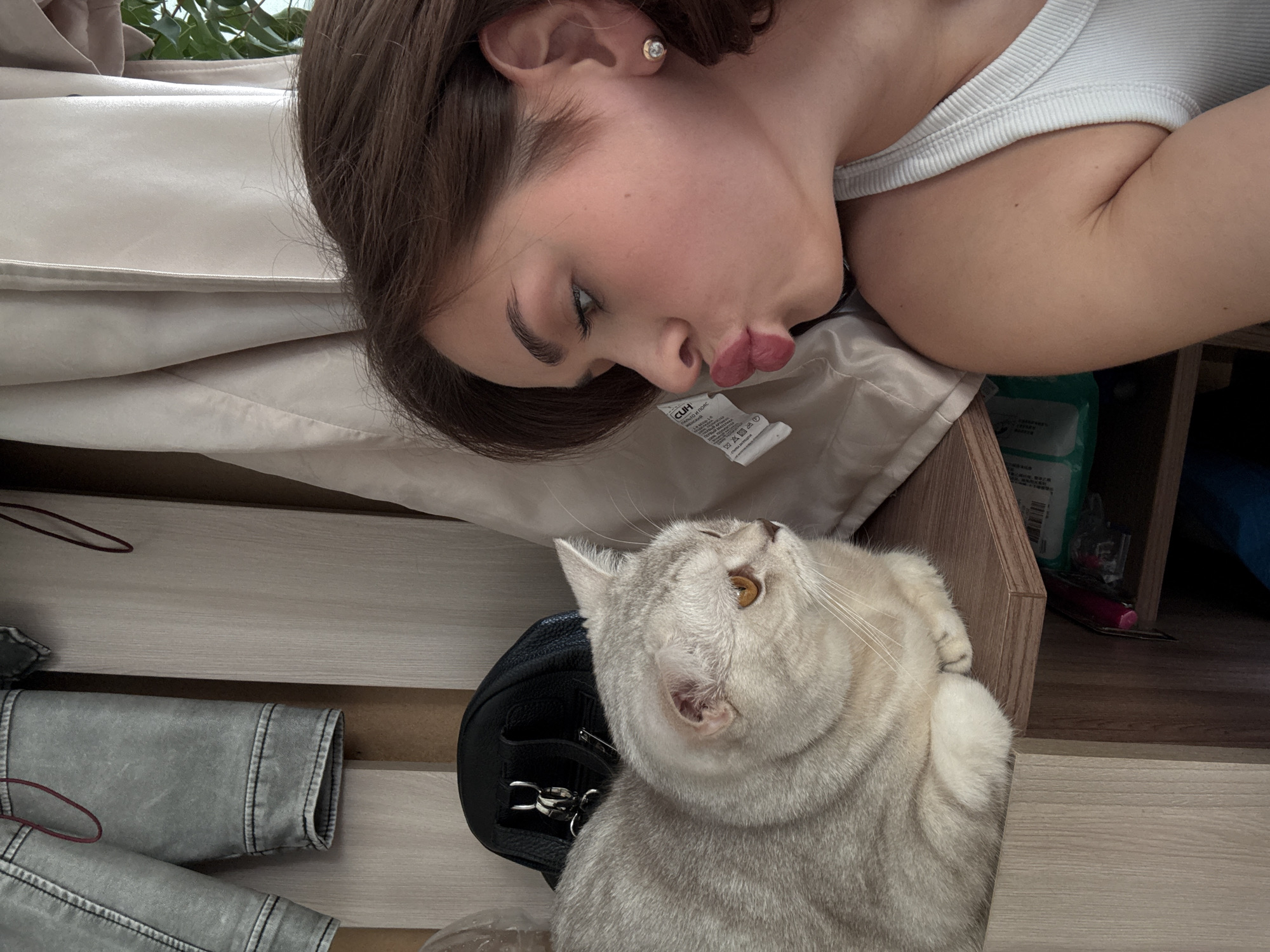 A medium shot depicts a light-skinned young woman, with dark brown hair, wearing a white tank top, lying in bed and puckering her lips towards a British Shorthair cat. The cat, with light silver-gray fur and amber eyes, is seated on a black purse. Both are positioned on top of what appears to be a wooden bed frame. A light gray denim jacket is visible in the lower left corner. To the right, beside the bed, are stacked items, some in green packaging. A tag is affixed to the bed sheet near the woman's face, with text in Russian. No indication is given that it is in Sarapul, Russia.