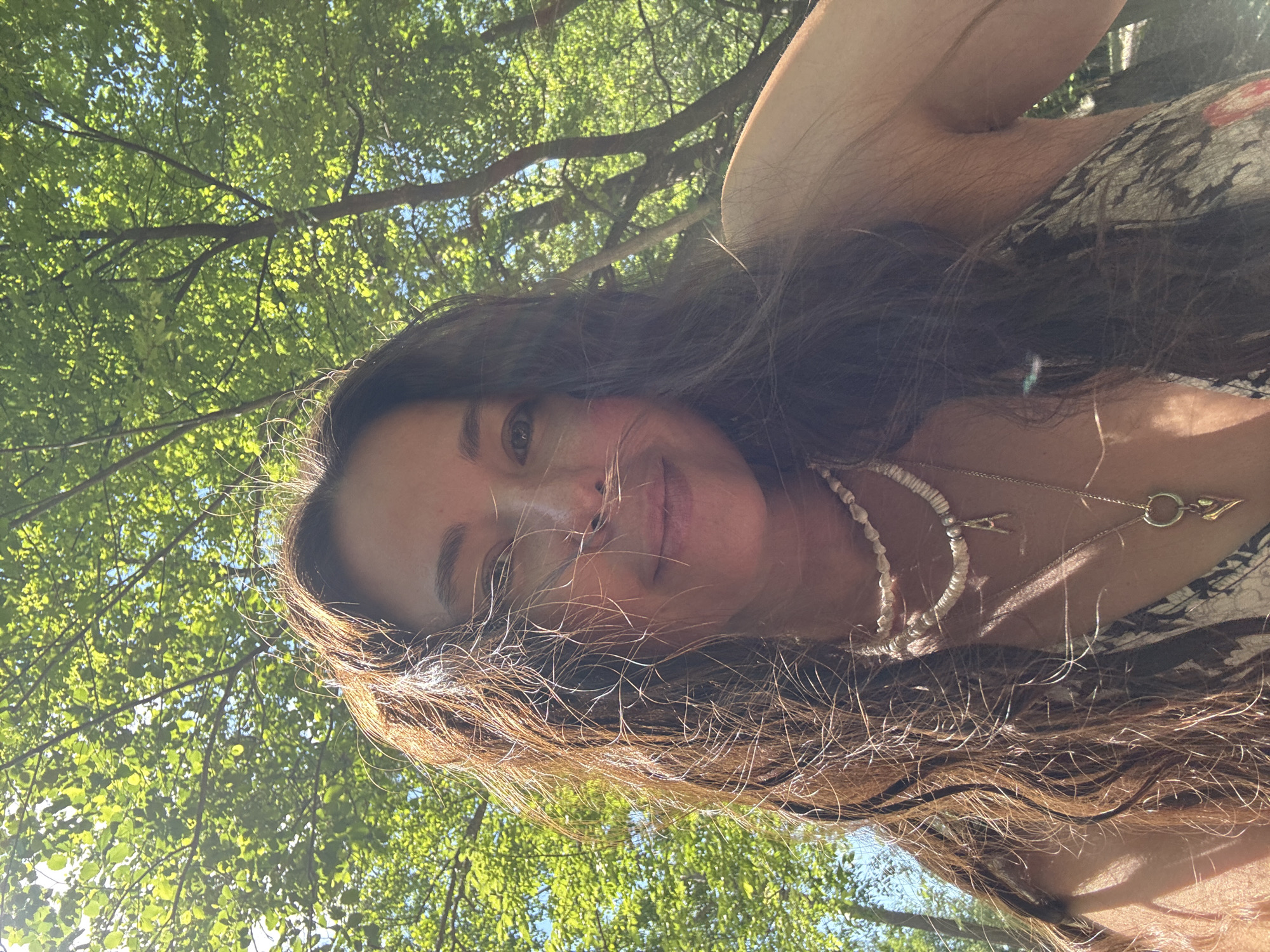 A woman with long, wavy brown hair smiles into the camera, her face partially obscured by her hair. She is wearing two necklaces: one with small beads and pearls, and another with a pendant shaped like a golden heart and an initial charm. She is outdoors, with lush green trees and sunlight filtering through the leaves in the background. The setting appears to be a park or a wooded area.