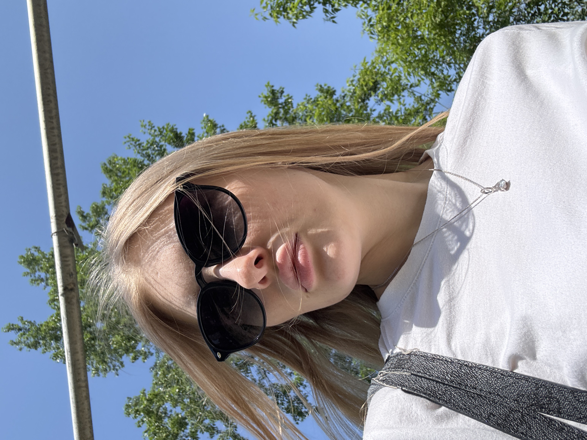 A medium shot, viewed from below, reveals a fair-skinned person with long, light brown hair and dark sunglasses. The person wears a white t-shirt and a silver necklace with a pendant. A dark grey woven strap is visible across the shirt. Behind the person, there are green trees and a clear blue sky. A gray metallic pole is positioned to the left. This image was taken outdoors, likely in Perm, Russia, judging from the location cues from associated images.