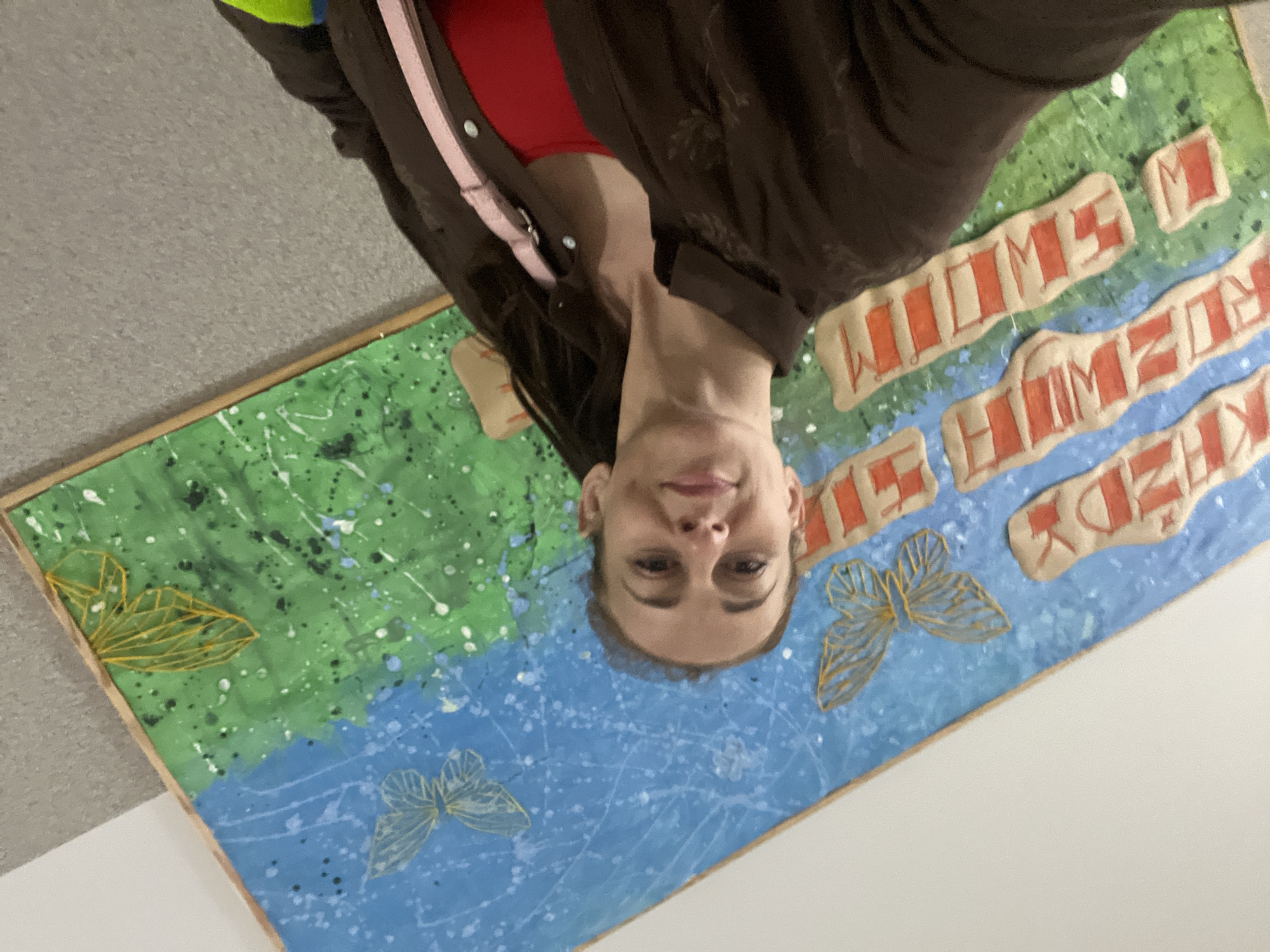A woman with brown hair and a brown button-up shirt over a red shirt takes a selfie in front of a large, colorful painting on a wall. The painting depicts a blue sky with white splatters and butterflies, and a green grassy field below with more butterflies. There are raised, textured letters on the painting that appear to be in an unfamiliar language. The woman is smiling and looking at the camera. The background is a plain wall.
