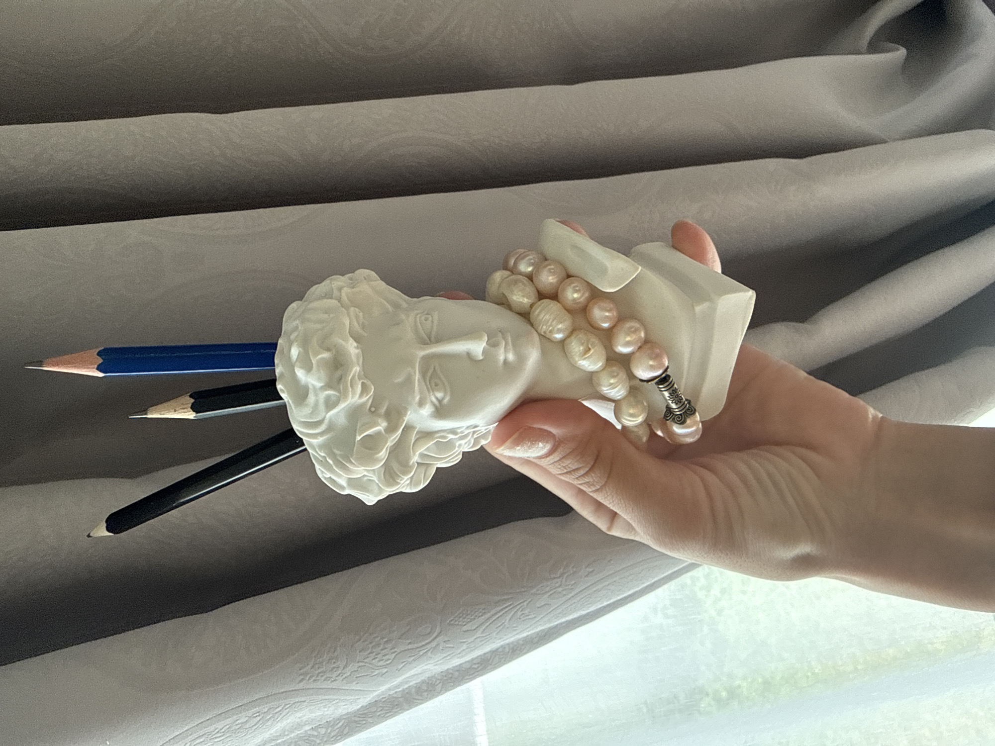A hand holds a white bust of a man, which functions as a pen holder. The bust is decorated with a pearl necklace. Three pencils are inserted into the top of the bust. The background consists of draped curtains. The scene appears to be indoors, near a window.