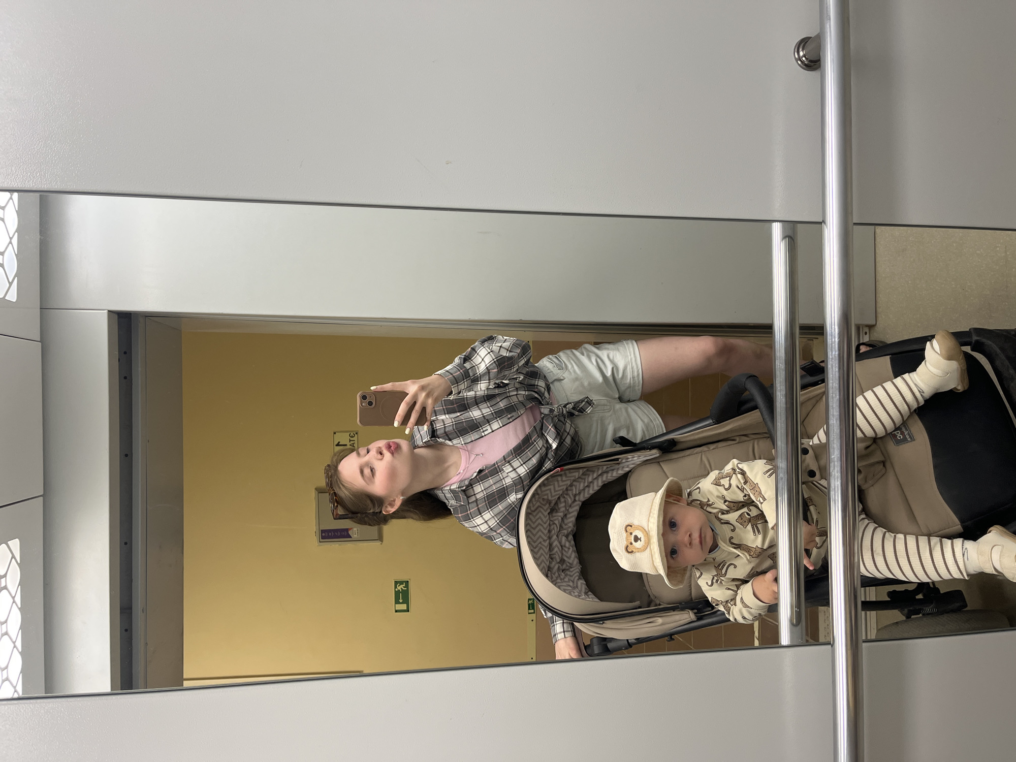 A woman is taking a mirror selfie with a baby in a stroller. The woman is wearing a pink shirt, a plaid jacket, and light-colored shorts. The baby is wearing a patterned outfit and a hat. The reflection shows they are in a room with beige walls and a white ceiling. There is a green exit sign on the wall. The location is Chaikovsky, Russia.