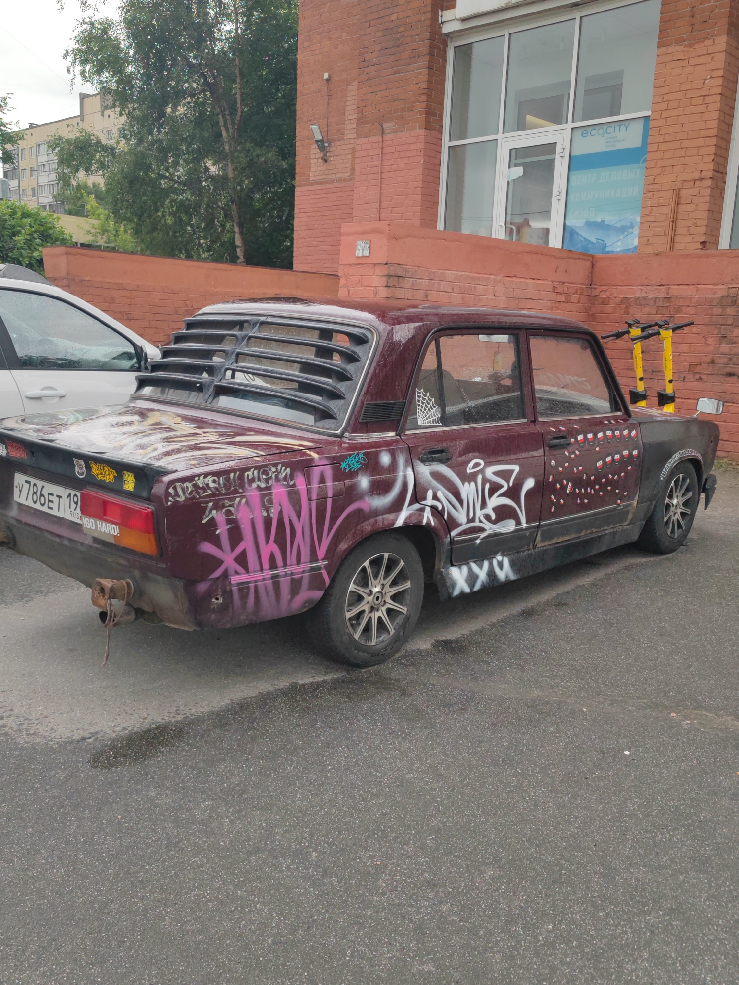 A maroon 1980s sedan, heavily customized with graffiti and stickers, is parked on an asphalt lot in front of a red brick building with large glass windows. The car has a custom rear window louvre and a tow hitch. Several yellow electric scooters are parked on the sidewalk behind the car. The building appears to be an office or commercial space, with a sign visible in one of the windows reading "ecocity" and "ЦЕНТР ДЕЛОВЫХ КОММУНИКАЦИЙ" (Center for Business Communications). The environment appears to be urban, likely in a residential or commercial district of Saint Petersburg, Russia, given the license plate which reads "Y786ET 19". The weather is overcast, and the asphalt is wet, suggesting recent rain. No people are visible in the image.