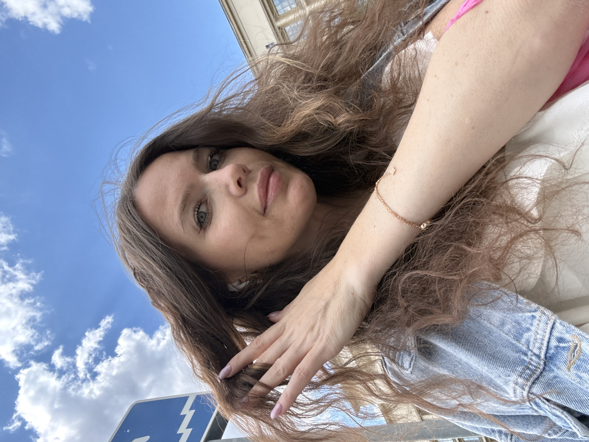 This is a bright, upward-angled selfie of a woman outdoors in Moscow, Russia, captured on a clear day. The primary subject is a young to middle-aged woman with long, flowing brown, wavy hair and blue-green eyes, looking directly at the camera with a neutral expression. She wears a light-colored denim jacket over a pink garment, and white wireless earbuds are visible in her ears. A delicate gold chain bracelet with a small rectangular charm adorns her right wrist, and her hand, seen near her hair, has light, possibly glittery pink nail polish.
The background features a vibrant blue sky with scattered white, fluffy clouds, indicating good weather and daytime. In the upper right corner, a part of an architectural building with windows is visible, suggesting an urban setting. A blue road sign with a white lightning bolt symbol is partially visible in the bottom left, further placing the scene in a city environment. The overall mood is casual and serene. No discernible text is visible beyond the lightning bolt symbol.