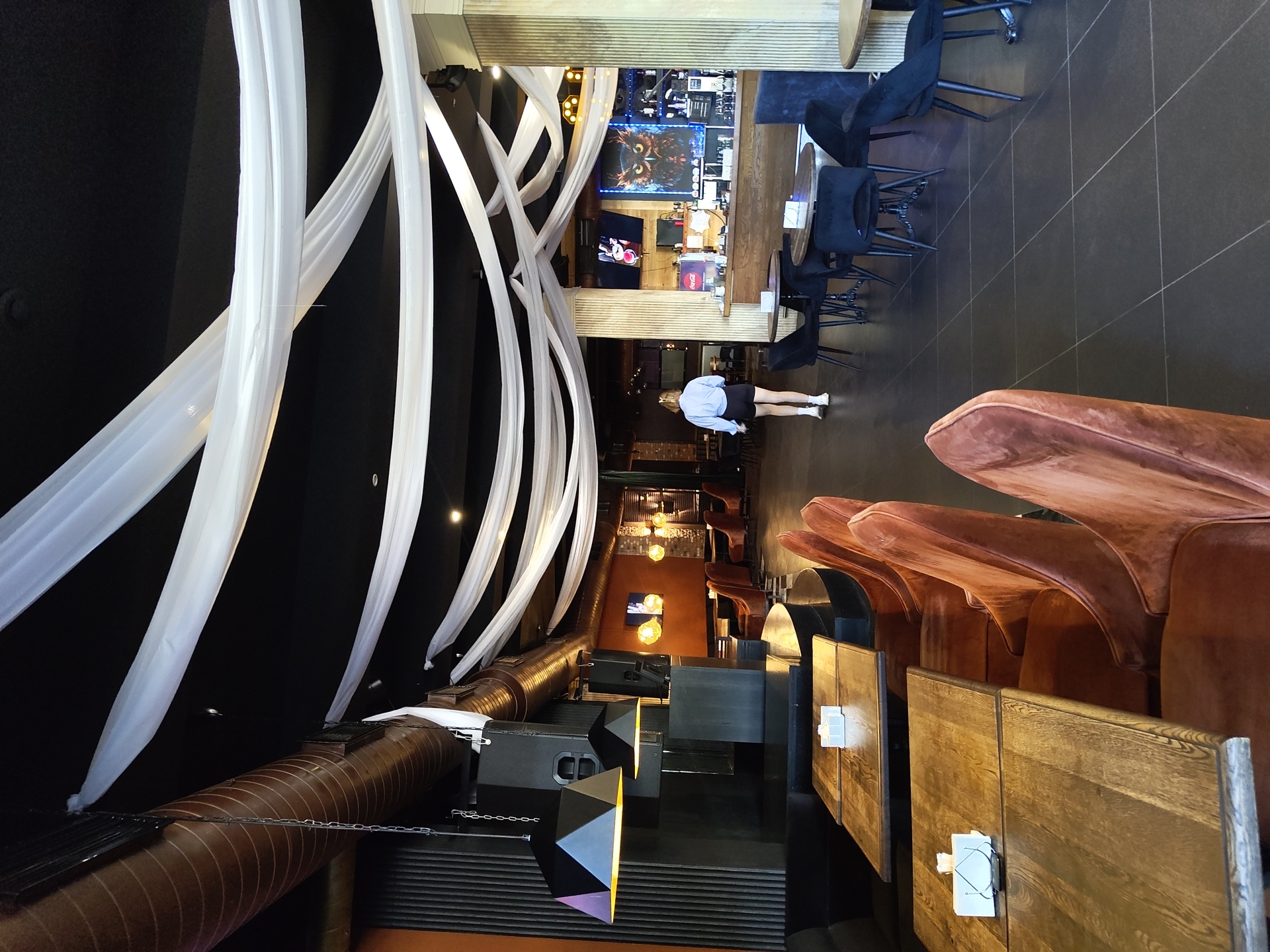 The image depicts the interior of a bar or restaurant in Zhukovsky, Russia. White fabric drapes are suspended from the dark ceiling, creating a decorative canopy. The space features several rows of wooden tables with chairs. In the foreground, plush, rust-colored velvet chairs are arranged around tables. Further back, black upholstered chairs surround round tables. A woman in a light blue shirt and black shorts is walking away from the camera towards the bar area. The bar itself has a wooden counter and is equipped with bottles and a large screen displaying an image of an owl. Exposed ductwork and industrial-style lighting are visible on the ceiling. The overall ambiance suggests a modern and somewhat upscale establishment.