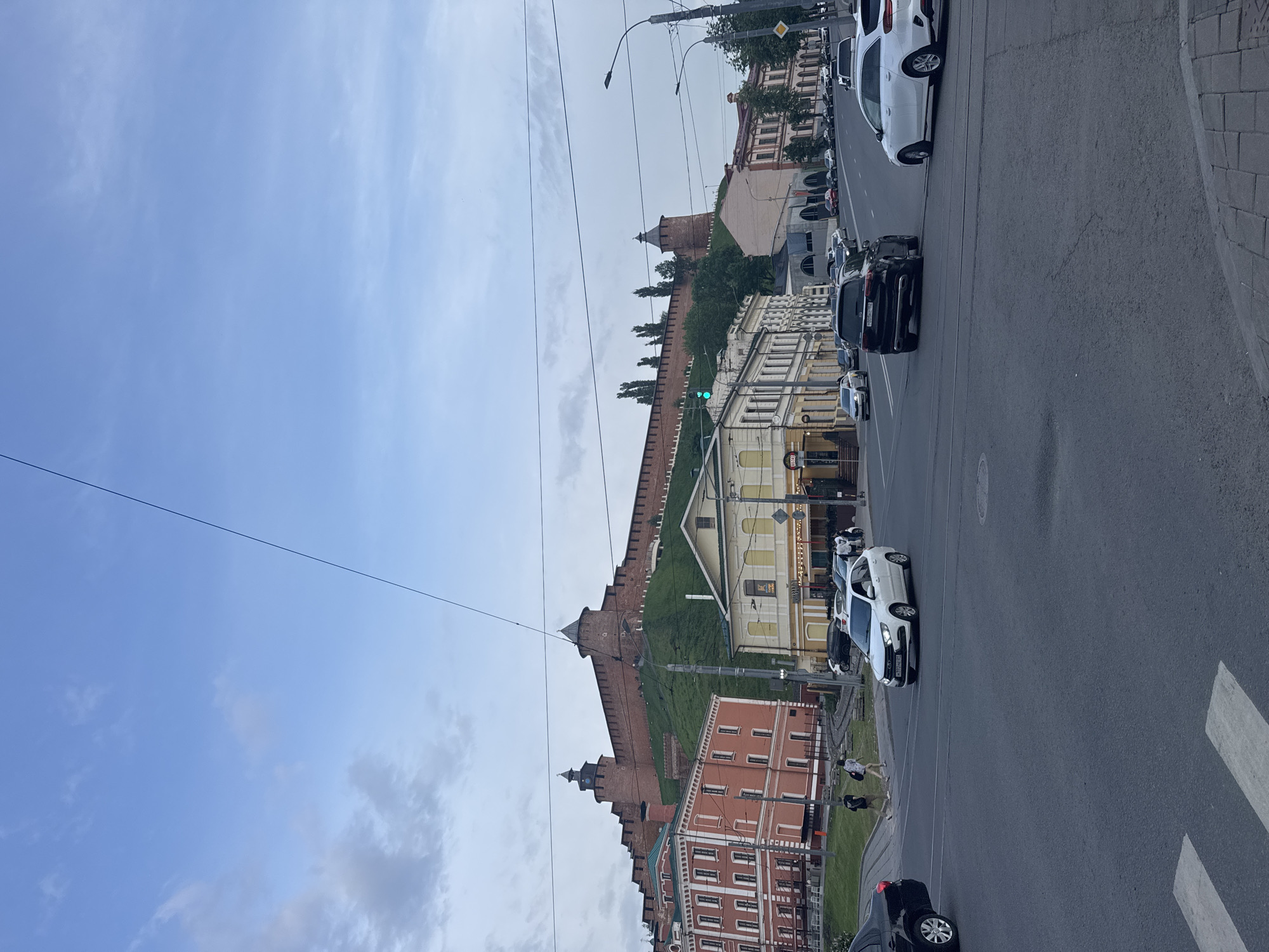 The image shows a street scene in Nizhny Novgorod, Russia. On the left, the walls and towers of the Nizhny Novgorod Kremlin are visible, rising up from a grassy slope. The Kremlin walls are constructed of red brick and have multiple towers. Along the street on the right are buildings of varying heights and colors, some of which appear to be commercial. Several cars are parked along the street and others are driving. A pedestrian crosswalk is visible in the foreground. Overhead are power lines and streetlights. The sky is overcast with patches of blue.