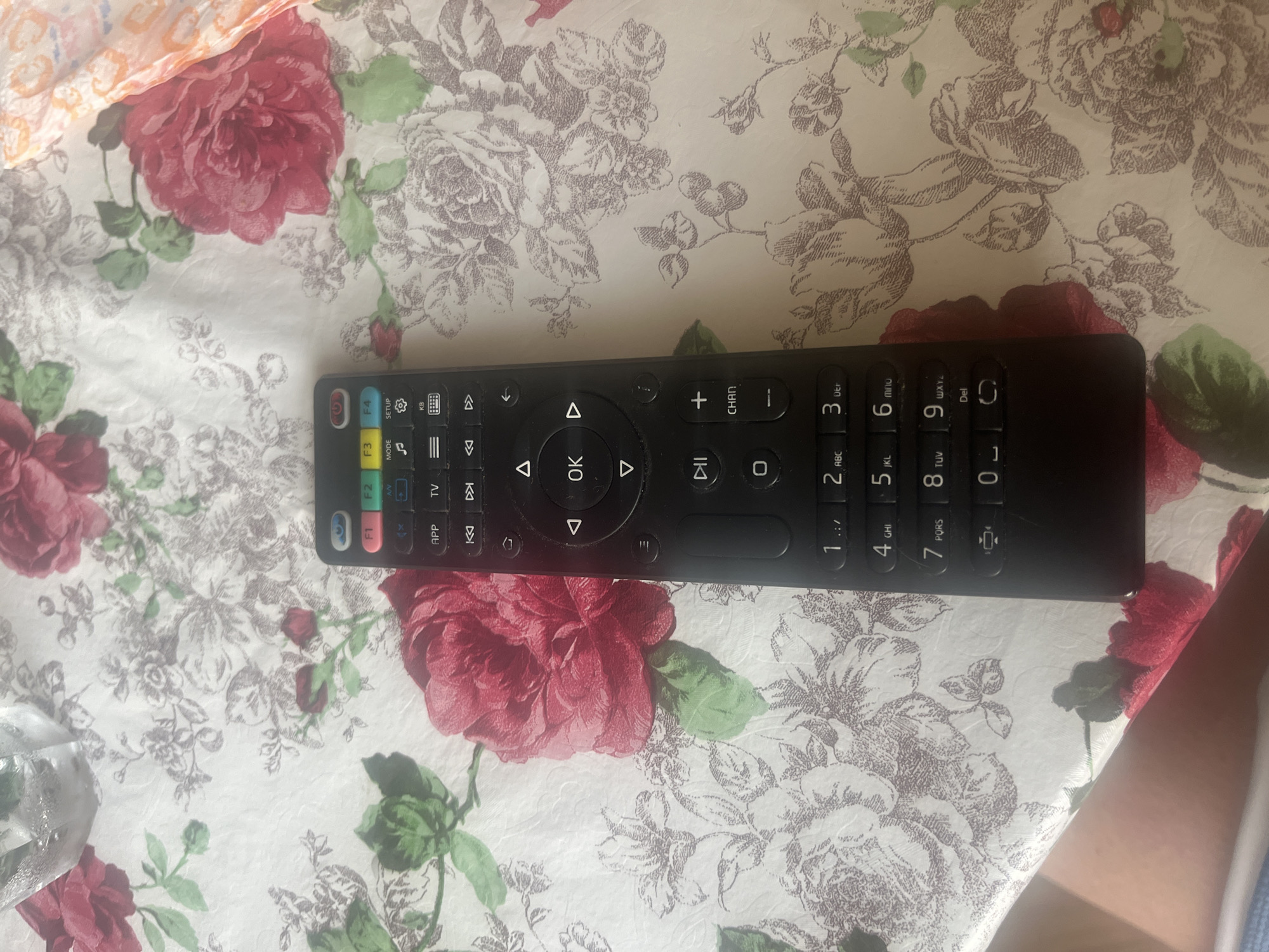 The scene depicts a tabletop in Burgas, Bulgaria, covered by a white floral tablecloth featuring a pattern of prominent red roses with green leaves and subtle greyish-brown floral sketches. A black television remote control is positioned vertically in the center-right of the frame, displaying various function buttons including colored F1-F4 buttons, power, mode, setup, mute, volume, channel navigation, media playback controls (rewind, play/pause, fast forward), a central directional pad with an 'OK' button, and a numeric keypad. In the upper-left corner, a clear glass containing a transparent liquid with visible bubbles is partially visible. The upper-right portion shows a crumpled piece of light-colored fabric with an orange geometric pattern. A light-skinned human forearm is visible in the lower-left quadrant, resting on the tablecloth, with a fragment of blue fabric (clothing) adjacent to it.