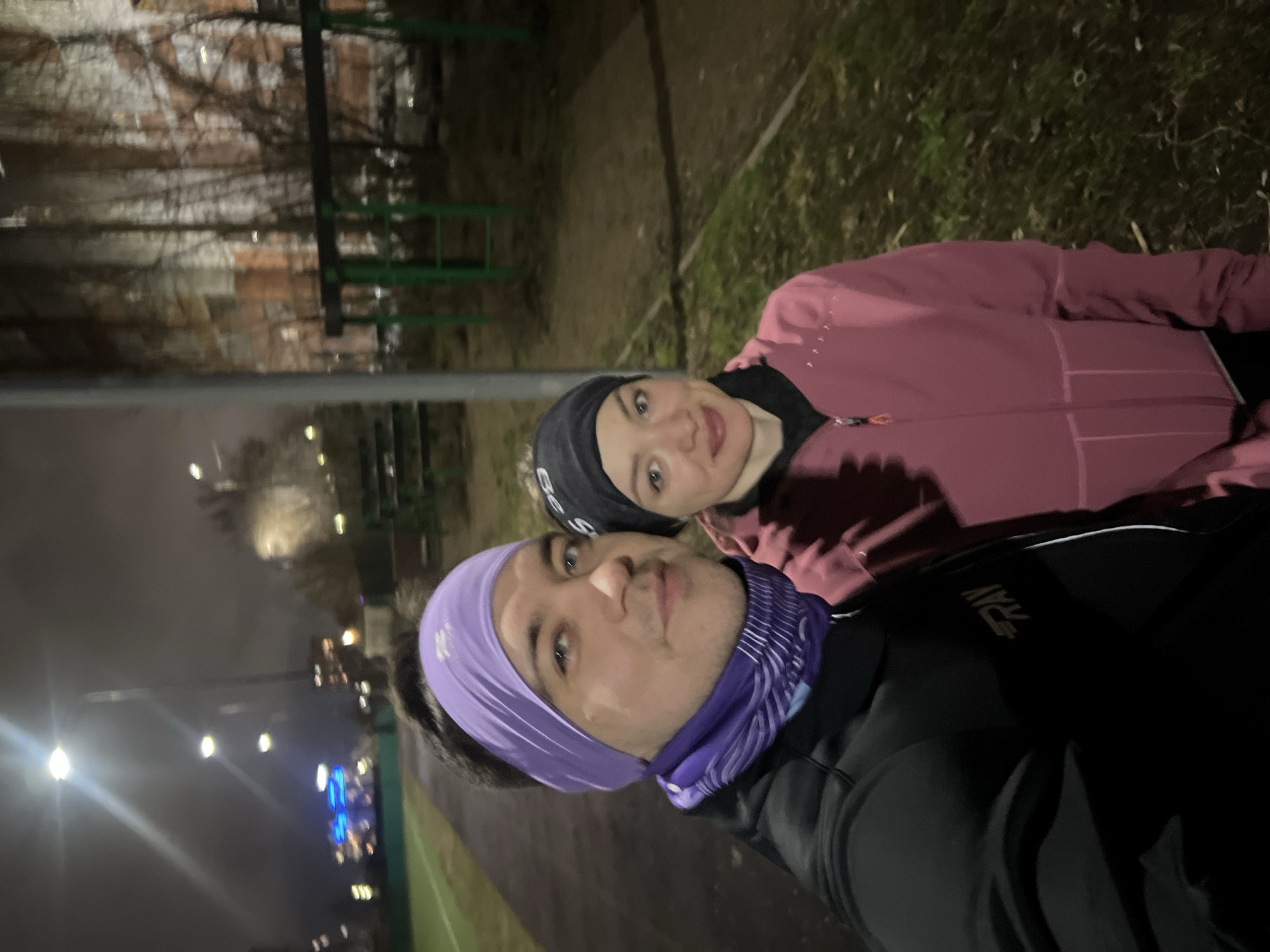 Two individuals are posing for a selfie in what appears to be an outdoor setting during the evening. The man in the foreground is wearing a black jacket, a purple headband, and a patterned scarf around his neck. The woman behind him is wearing a pink jacket and a black headband. The background shows some vegetation, possibly a park or grassy area, and a building with lit windows in the distance. Based on the available information, it's challenging to confirm the exact location within Kazan, Russia, without more visual cues.