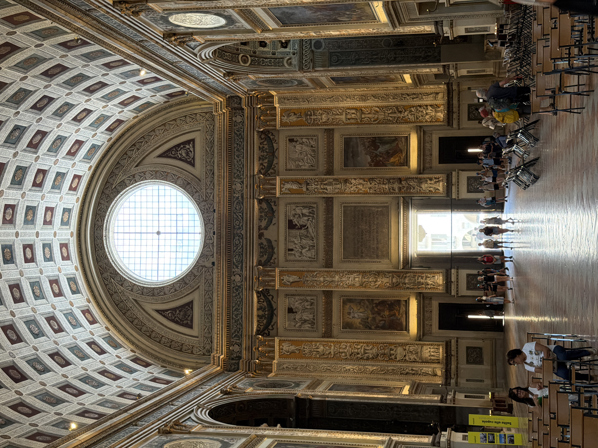 The image shows the interior of the Basilica di Sant'Andrea in Mantua, Italy. The ornate coffered dome and the oculus that provides natural light are the dominant features. The walls are decorated with paintings, sculptures, and intricate architectural detailing. Several people are present: some are seated in rows of chairs, while others are walking toward a brightly lit doorway in the background. Informational posters are visible in the foreground.