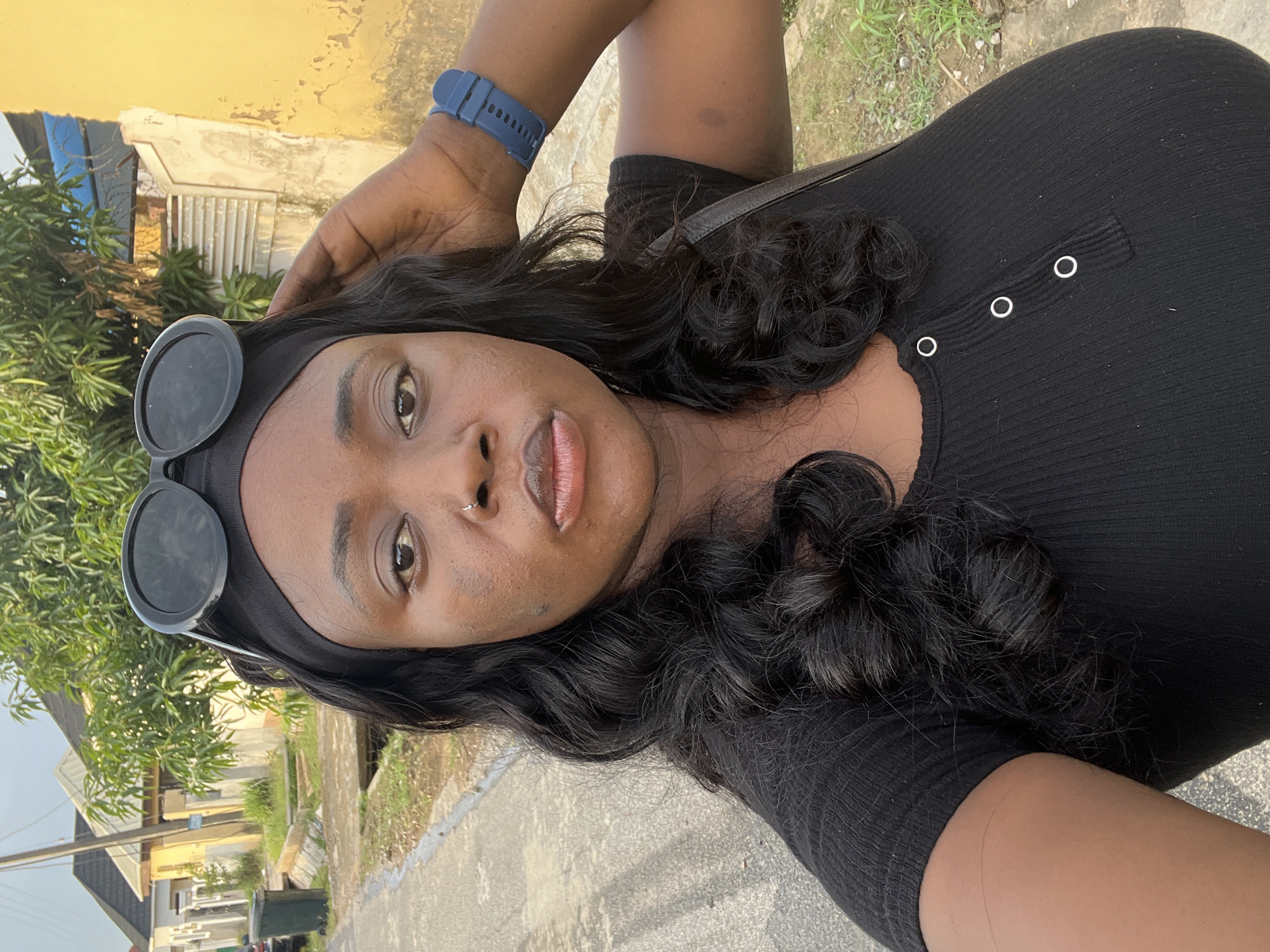 A medium, upward-angle, and rotated close-up shot of a Black woman standing on what appears to be a paved residential street in Abuja, Nigeria. The woman has dark hair styled in loose waves, with sunglasses pushed up on her head. She is wearing a black, fitted top with three silver buttons at the neckline. A blue smartwatch is visible on her wrist.
In the background, there is a yellow building, trees, a section of a white house, and a street. The lighting suggests it is daytime.