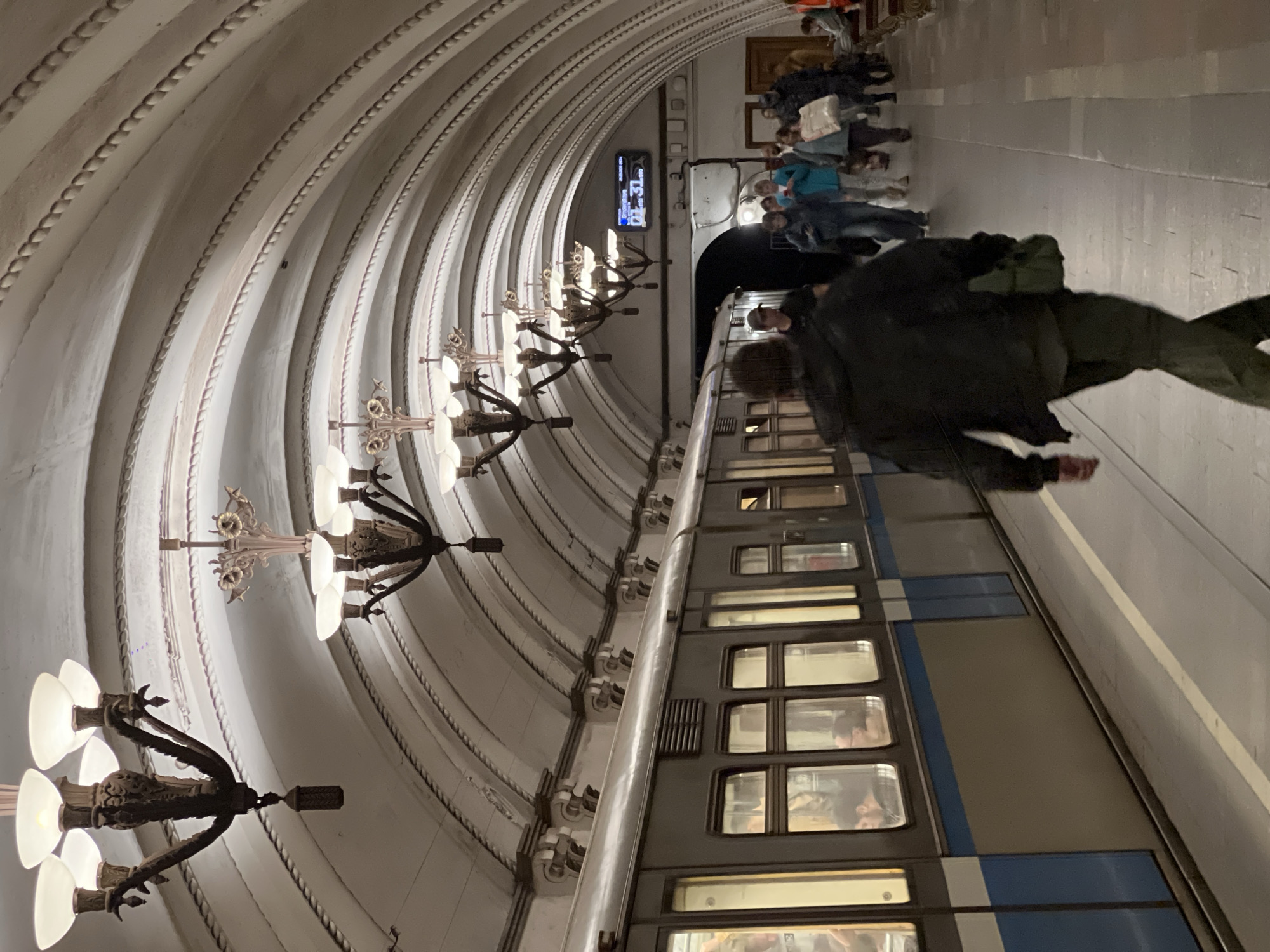 The image shows a subway station in an unspecified location. The arched ceiling of the station features decorative ribs and ornate chandeliers. A subway train is alongside the platform, with people visible inside the train cars and standing on the platform. A digital display shows the time.