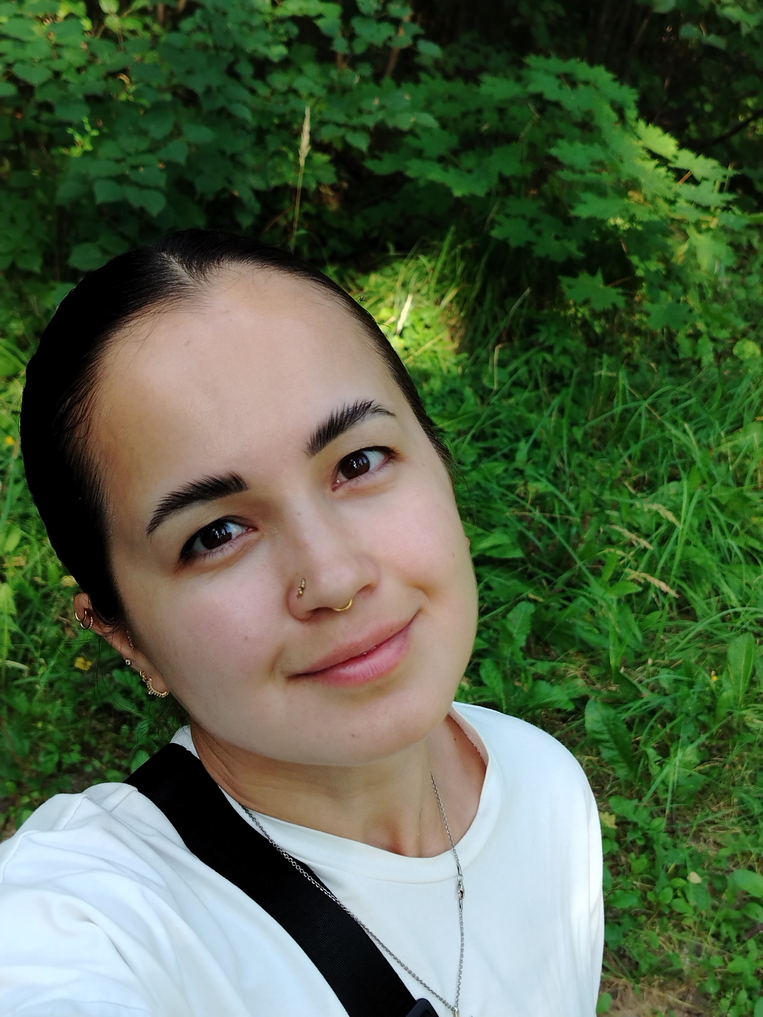A close-up, high-angle selfie is captured of a person with dark hair pulled back, a nose ring, and ear piercings, wearing a white t-shirt. The person is smiling slightly, with their head tilted. The background consists of lush green foliage, indicative of an outdoor, natural setting, possibly in the city of Noviy, Russia. The lighting suggests it is daytime, with natural sunlight filtering through the trees.