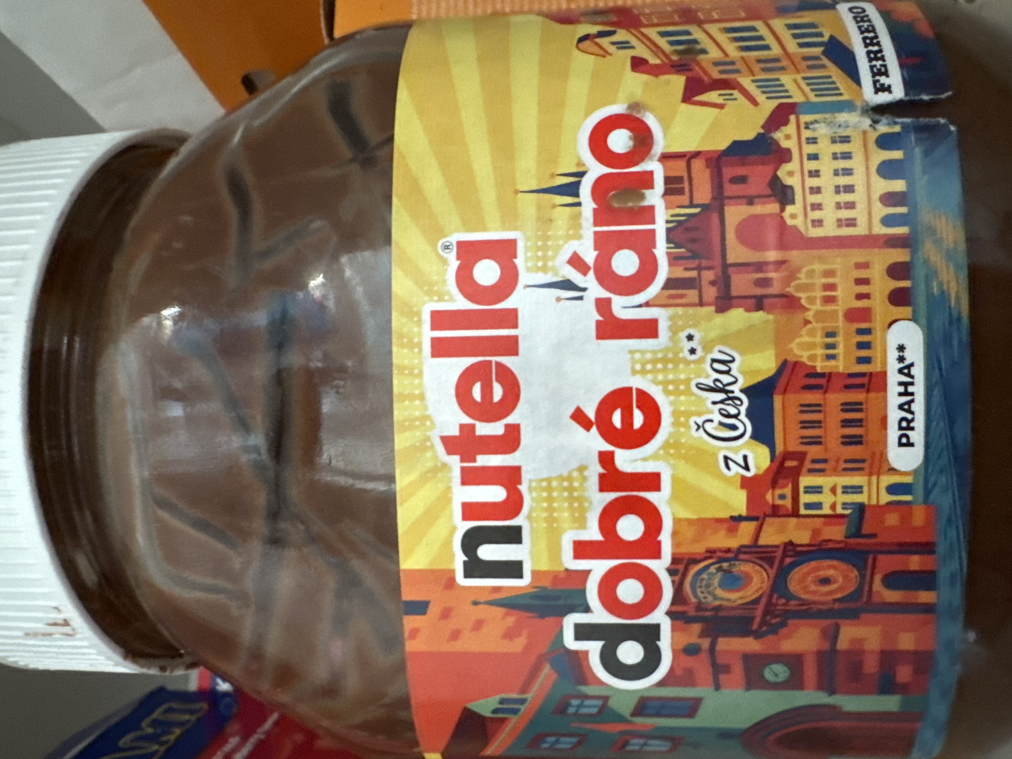 A close-up, slightly angled shot shows the label of a jar of Nutella. The label is predominantly yellow with a colorful illustration of a European city skyline, featuring buildings with red roofs, arched windows, and a prominent clock tower. The word "nutella" is displayed prominently in large red and white letters across the top of the label. Below it, in larger red letters, is the phrase "dobré ráno" with a small "z Ceska" written below and to the right of "dobré." At the bottom of the label, a white oval shape contains the text "PRAHA**" and to the right, the word "FERRERO" is printed in white on a black background. The jar itself appears to be made of brown glass, containing the dark brown hazelnut spread. The lid is white. The background is blurred, suggesting the jar is on a shelf or table, with hints of other packaging and possibly a wall or shelf. There is no indication of the location being in Agios Tychona Community, Cyprus; the visible text suggests a connection to Prague, Czech Republic.