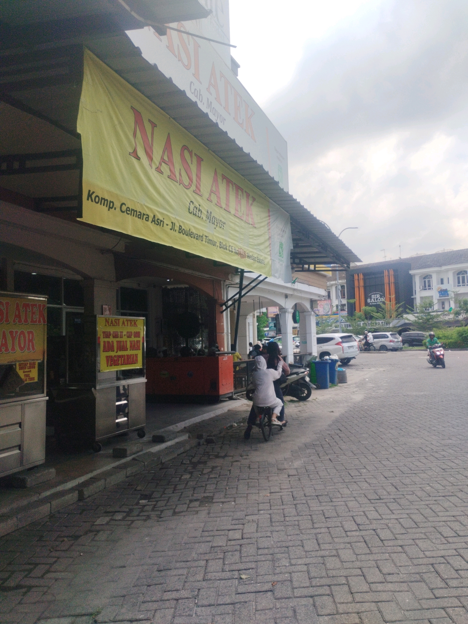 The image depicts an outdoor scene in Percut Sei Tuan, Indonesia, featuring a food stall area and a street. Dominating the left side is a yellow banner with red lettering that reads "NASI ATEK" and "Komp. Cemara Asri - Jl. Boulevard Timur, Blok CS". Below this, another banner or sign is visible with text including "SI ATEK" and "AYOR". To the right of the food stall, a smaller yellow sign with red and black text reads "NASI ATEK / YUP GIRI II - CAP GOM / ADA JUAL NASI VEGETARIAN".
In front of the food stalls, there are several metallic display cases and what appears to be a generator or large appliance in orange. People are present in the scene; on the left, a person is seated on a bicycle, facing away from the camera, dressed in white. To the right of the food stall, two people are on a motorcycle, one driving and the other as a passenger, both facing away. Further down the street, another person is riding a motorcycle and another motorcycle is parked.
The environment includes a paved sidewalk or walkway made of interlocking grey bricks, which extends into a street. The background reveals buildings, including a modern-looking structure with "SIR SALON" signage and other residential or commercial buildings with white facades. The sky is overcast, suggesting a cloudy day. The overall scene suggests a busy commercial area with food vendors and street activity.