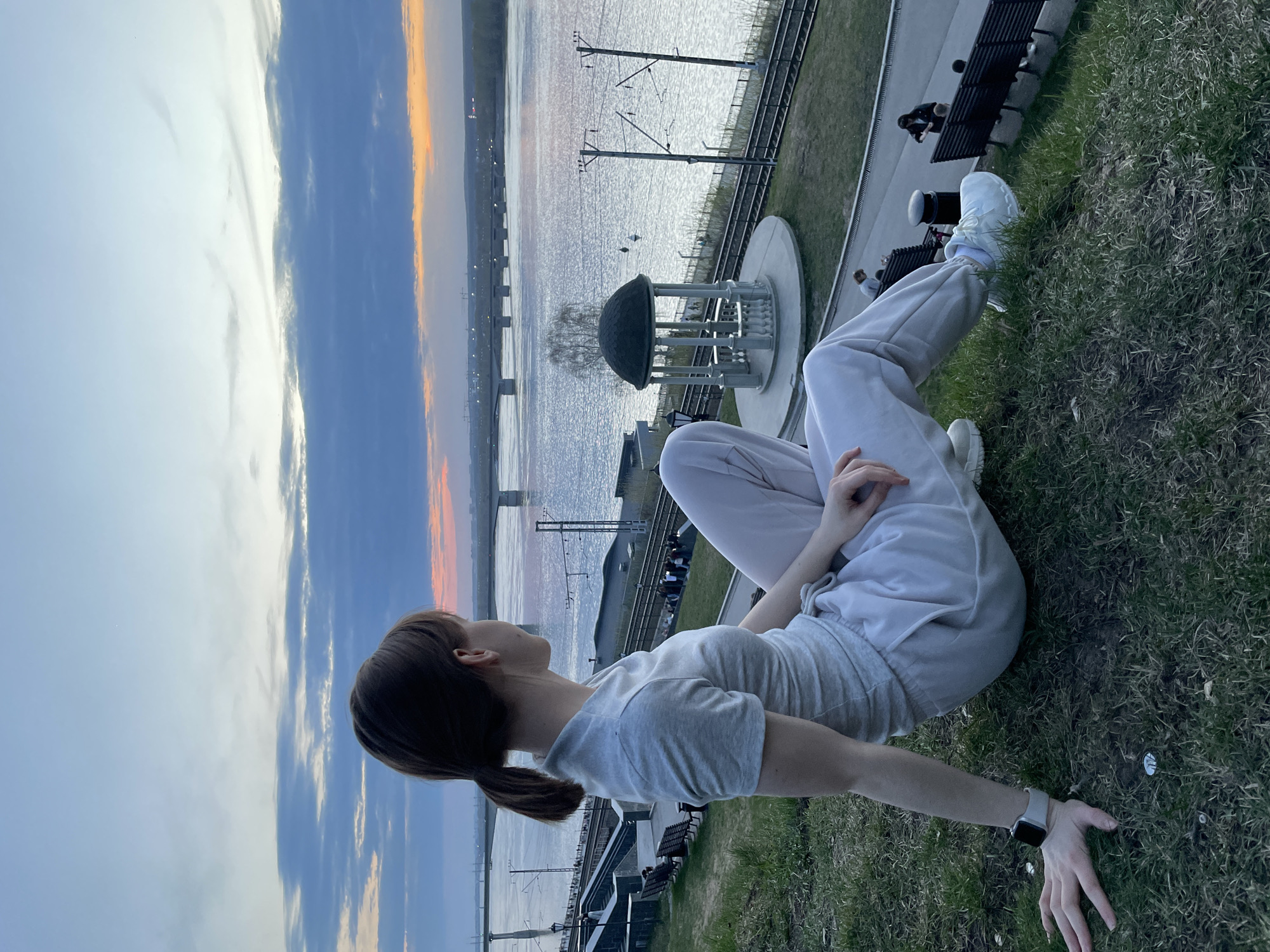 A woman sits on a grassy slope overlooking the Kama River in Perm, Russia. She wears a gray t-shirt, light gray sweatpants, and white sneakers. Her dark hair is tied back in a ponytail, and she has a smartwatch on her left wrist.
In the background, the Kama River reflects the sky at sunset, with a bridge visible in the distance. An observation point and a railway line run along the riverbank. Benches and people are seen along the walkway.