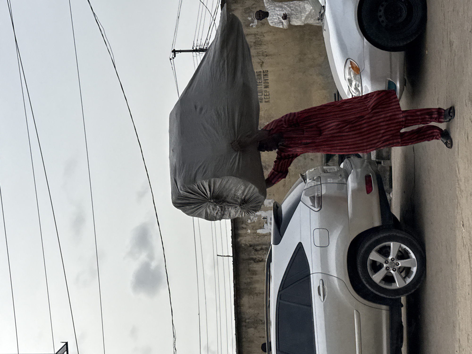 This image depicts an outdoor street scene in Lekki, Nigeria, under an overcast sky. The central focus is a man, dressed in a red and black striped long-sleeved shirt and trousers, who is actively moving or lifting a large, irregularly shaped bundle wrapped in grey sack-like material. He grips the bundle with both hands, appearing to exert significant effort.
Behind the main subject, a light-colored, worn concrete or plastered wall features white painted text that clearly reads: "NO LOITERING KEEP MOVING". Another individual, wearing a light-colored garment, is partially visible standing near the wall and the front section of a silver sedan. Further to the right, the rear part of a silver SUV is also visible, parked on the street. Overhead, numerous power lines or utility cables stretch across the muted sky. The scene captures a moment of activity, likely involving the transport of goods, within an urban or semi-urban environment.