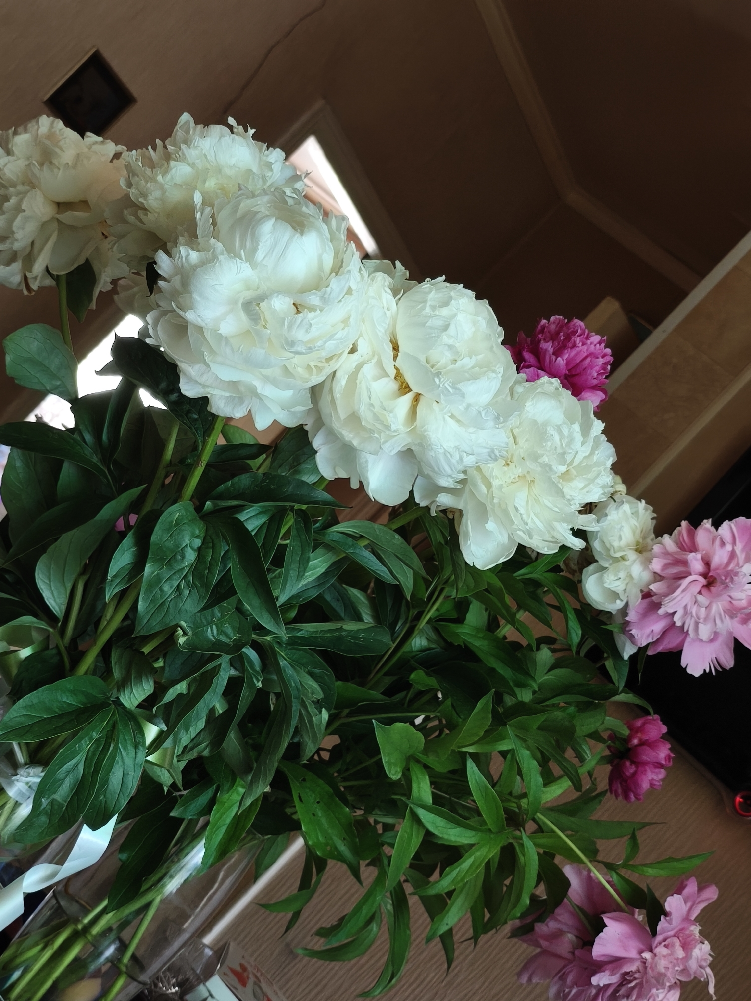 A bouquet of peonies is displayed in a clear glass vase. The peonies are a mix of white, pink, and magenta. The white peonies are large and full, with ruffled petals. The pink and magenta peonies are smaller and more compact. The bouquet also includes dark green leaves. The vase is placed on a light brown wooden surface. In the background, a wall with a textured surface is visible, and a dark object, possibly a television screen, is partially visible to the right. The lighting suggests an indoor environment, likely a home or a display area.