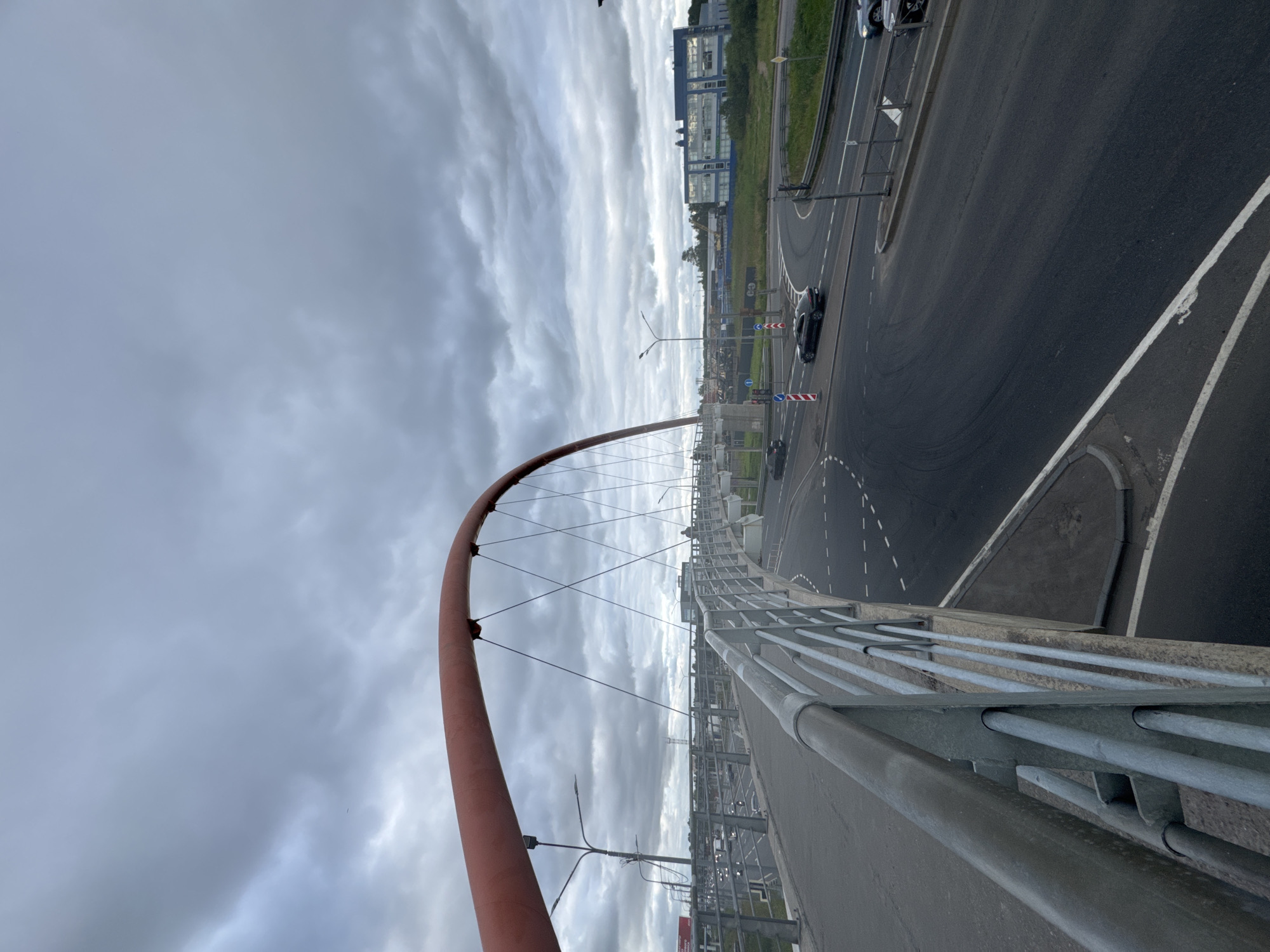 The image shows a pedestrian bridge in Saint Petersburg, Russia, over a busy road junction. The bridge has a red arched structure and a metal railing. Several cars are visible on the road below, some navigating a roundabout. In the background, there are modern buildings and overcast skies. The scene is captured from the perspective of someone standing on the pedestrian bridge, looking downwards and across the road.
