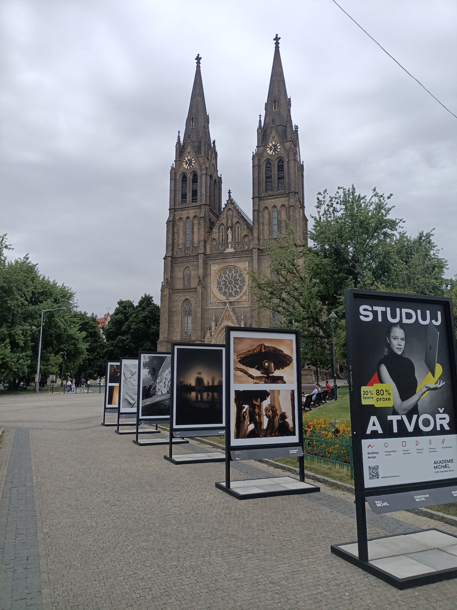 The image depicts an outdoor art exhibition in front of the Basilica of St. Peter and St. Paul in Vyšehrad, Prague, Czechia. The basilica, a prominent Neo-Gothic structure with two spires and a large rose window, is the dominant background element under an overcast sky. In the foreground, a row of framed photographs is displayed on black stands. These photographs appear to be a mix of black and white and color images, some featuring abstract or artistic subjects, while others depict what might be sculptures or figures.
To the right of the photographs, a large black advertisement board is visible. It features the word "STUDUJ" (Study) in large white letters at the top, followed by an image of a young woman holding a laptop. Below this, text in Czech, including "20% teorie 80% praxe" (20% theory 80% practice) and "A TVOŘ" (And Create), is displayed. Further down, the advertisement lists various fields of study such as "Grafický design" (Graphic Design), "Fotografická tvorba" (Photographic Creation), "Design interiérů" (Interior Design), "Filmová tvorba" (Film Creation), and "Scenáristika" (Screenwriting), along with "AKADEMIE MICHÄEL" and a QR code.
On the left side of the image, a paved walkway leads towards the basilica, with a grassy area and trees separating the exhibition from the building. Several people are visible in the background, walking along the path or sitting on benches in the grassy area. The overall scene suggests an event or exhibition taking place in a public, culturally significant location within Prague.