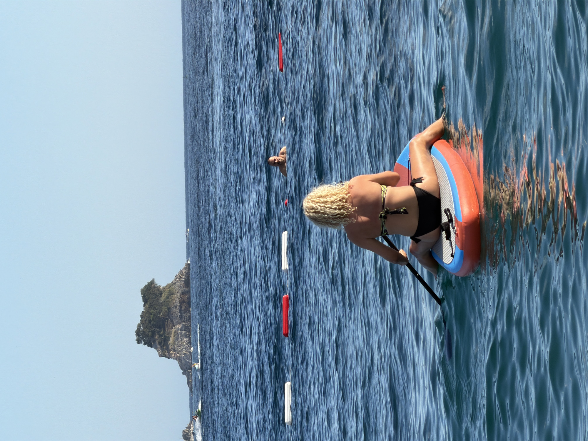 This is a photograph taken from the water in Budva, Montenegro, showing a woman on a paddleboard. She is facing away from the camera, with her blonde, curly hair visible. She is wearing a black bikini and is holding a paddle. The paddleboard is orange and blue. In the background, there is a small island covered in trees, likely Sveti Nikola Island, which is a prominent landmark off the coast of Budva. There are also jet skis and other boats in the distance, suggesting a popular recreational area. The water is a deep blue and appears somewhat choppy. Three buoys, two white and one red, are visible in a line across the water. A man is partially visible in the water behind the paddleboarder, further out in the sea. The sky is clear and blue.