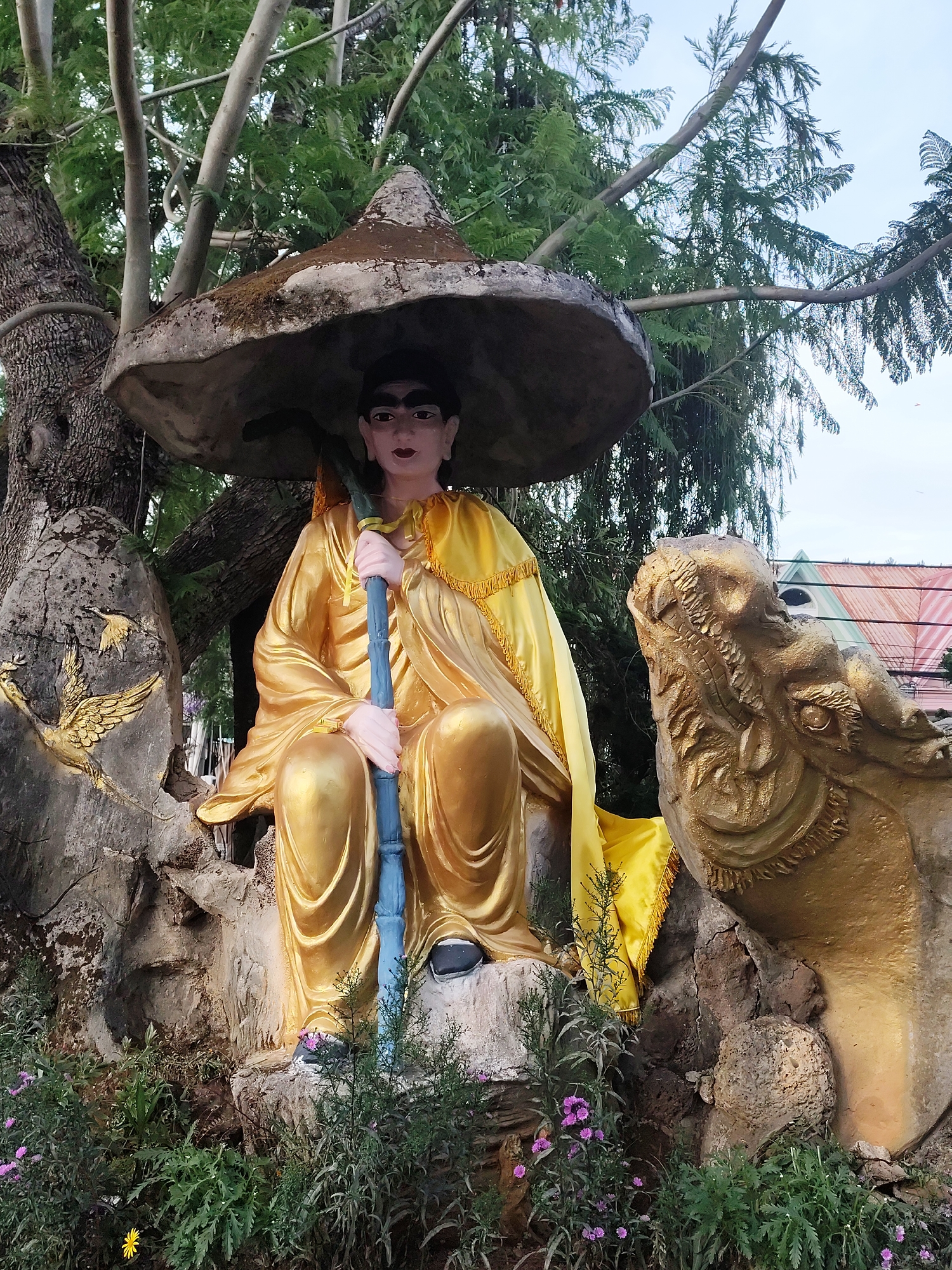 This image shows a statue of a seated figure in a seated position, situated within an outdoor setting, likely a garden or park. The location is in Cam Ly - Đà Lạt, Vietnam.
The figure is dressed in golden robes and wears a large, conical hat that shades the head and shoulders. The face is painted with defined features, including eyebrows and lips. The figure holds a blue pole or staff, and the overall pose appears contemplative.
The statue rests on what appears to be a rock-like base, with surrounding vegetation including purple flowers. To the statue’s right is a representation of a golden dragon, with textured scales and prominent features.
The statue is positioned underneath a large tree, which extends to the sky. The surroundings suggest a temple or garden, located in a city (Cam Ly - Đà Lạt) in Vietnam.